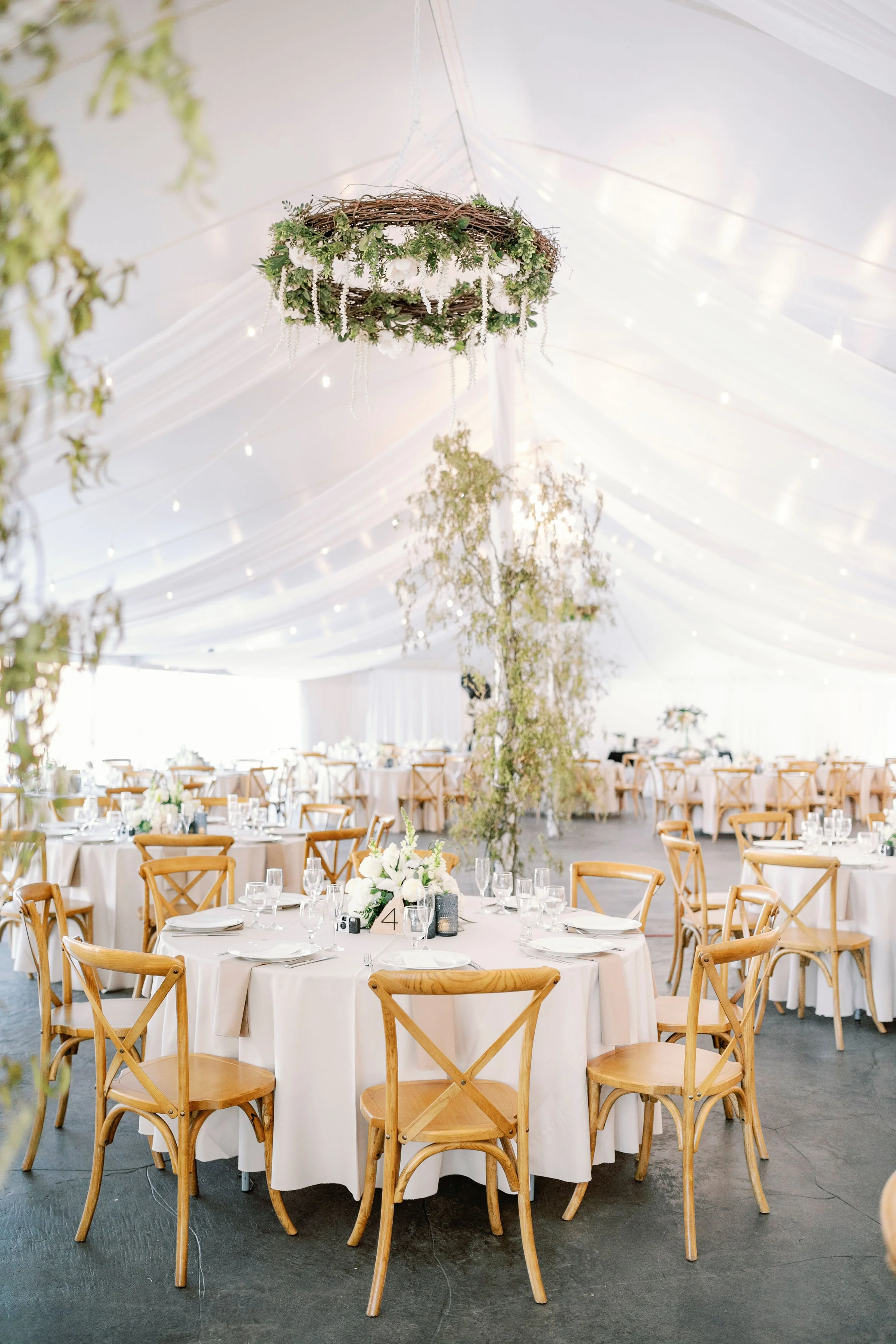 Wedding reception setup in a white tent with round tables, wooden chairs, floral centerpieces, and hanging decorations.
