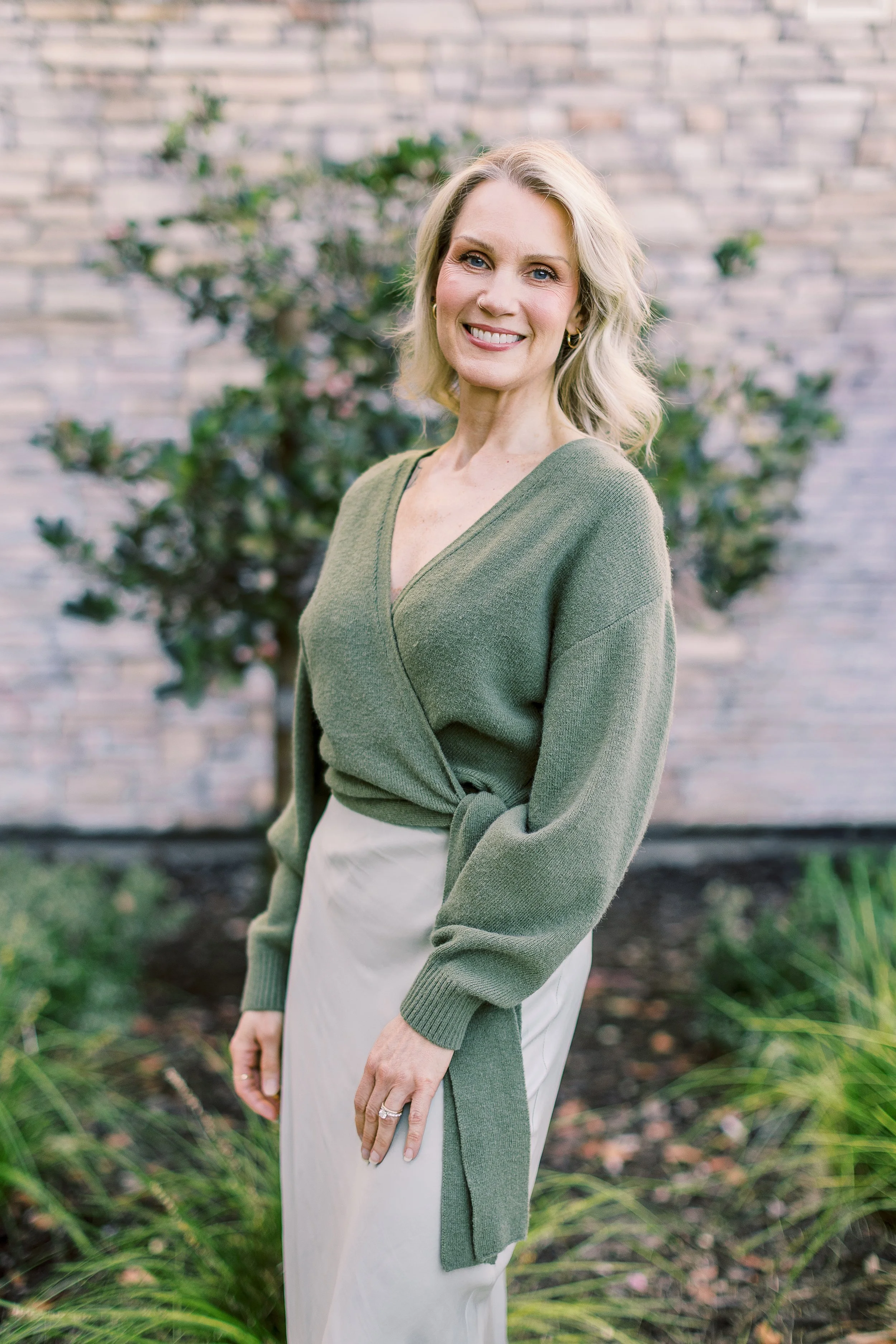 A woman with blonde hair and a smile, wearing a green wrap sweater and a light-colored skirt, standing outdoors with greenery and a brick wall in the background.