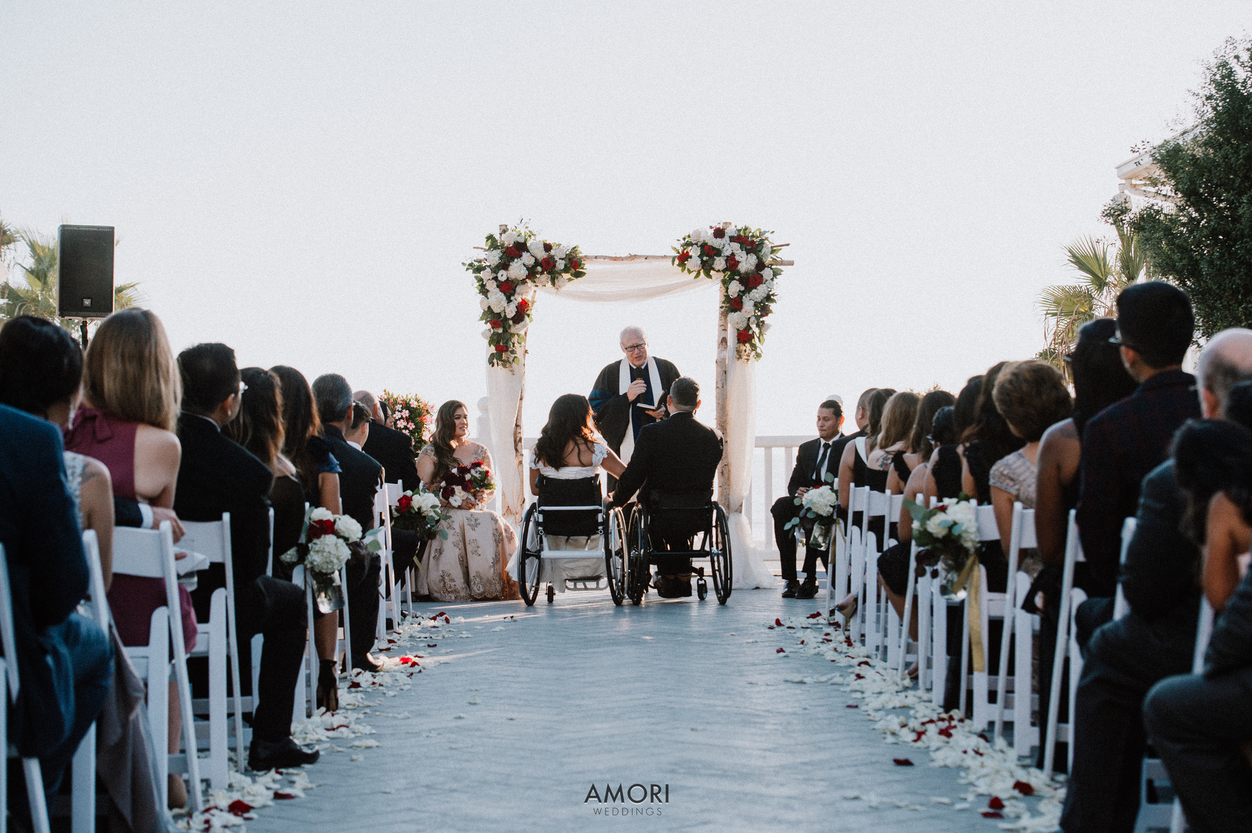 A wedding ceremony with a diverse group of guests, some in wheelchairs, seated on either side of a decorated aisle with rose petals. The officiant is standing under a floral arch, conducting the ceremony outdoors during daytime.