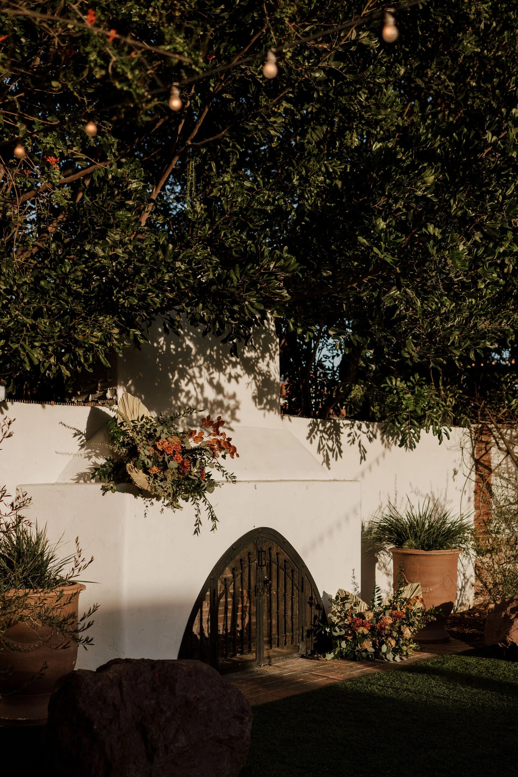 Decorative outdoor fireplace with flower arrangements and potted plants, surrounded by greenery and illuminated by warm sunlight.