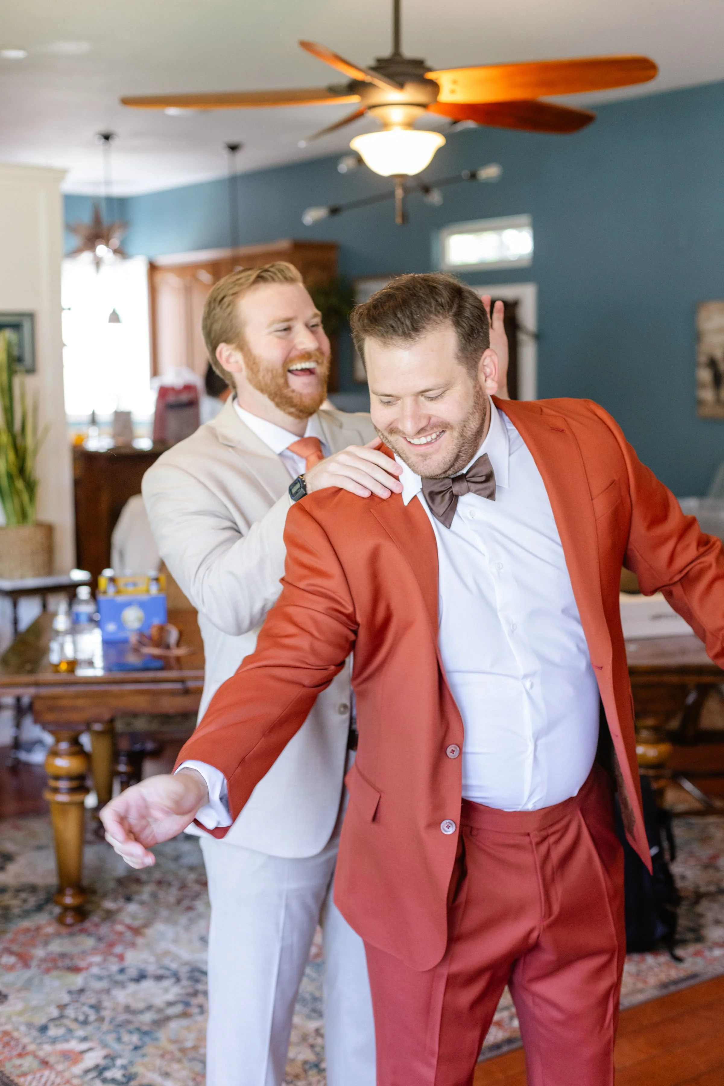 Two men dressed in suits, one in a beige suit and the other in a red suit with a bow tie, laughing and sharing a joyful moment in a warmly decorated living room.
