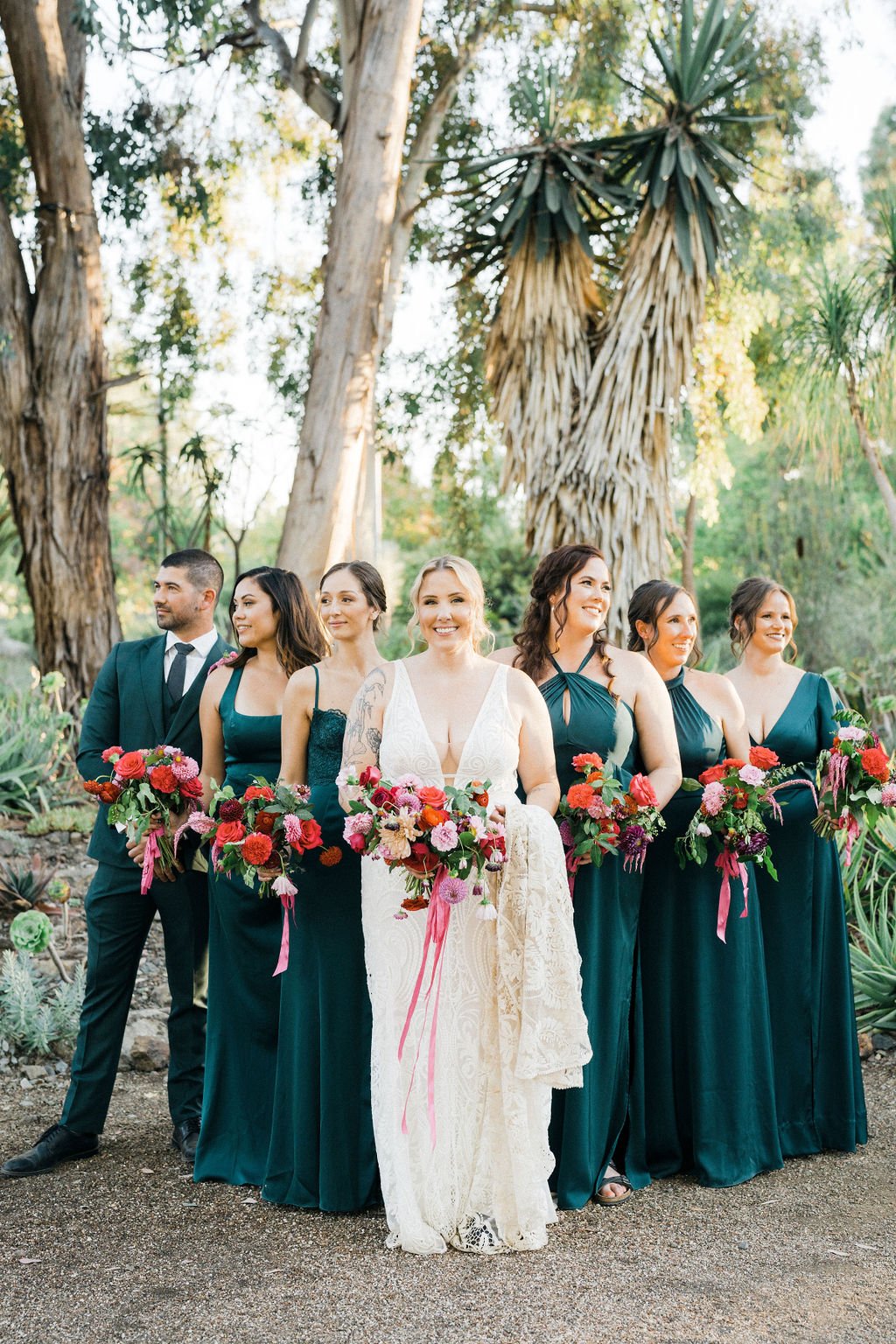 A bridal party of six women and one man stands outdoors among trees, holding colorful bouquets of flowers. The bride, in a white wedding dress, is in the center, smiling, with her bridesmaids in dark green dresses on either side. A groomsman in a suit stands on the far left.
