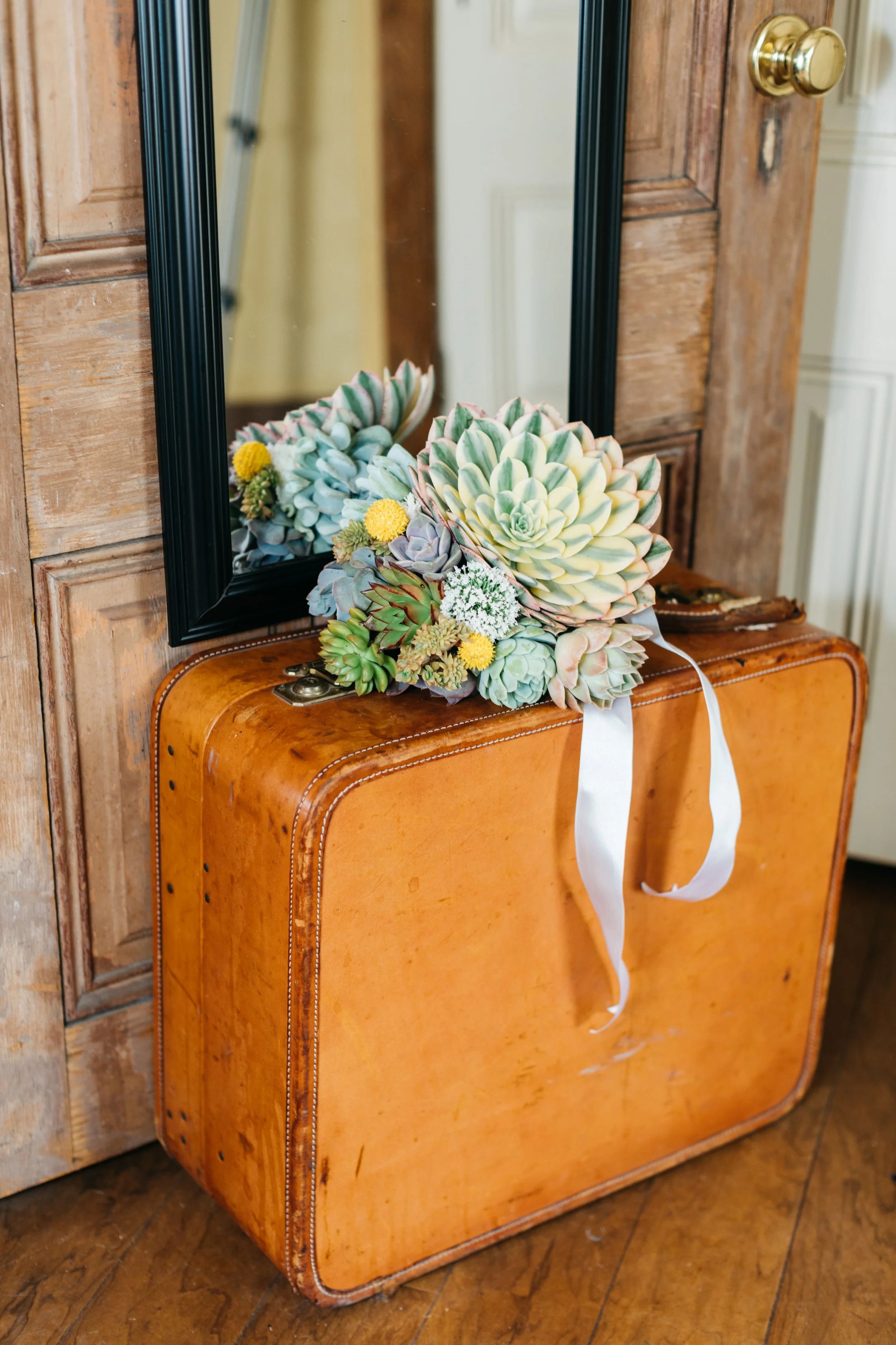 A mirror reflecting a bouquet of succulent plants and yellow flowers resting on an orange vintage suitcase in front of a wooden door with a brass doorknob.