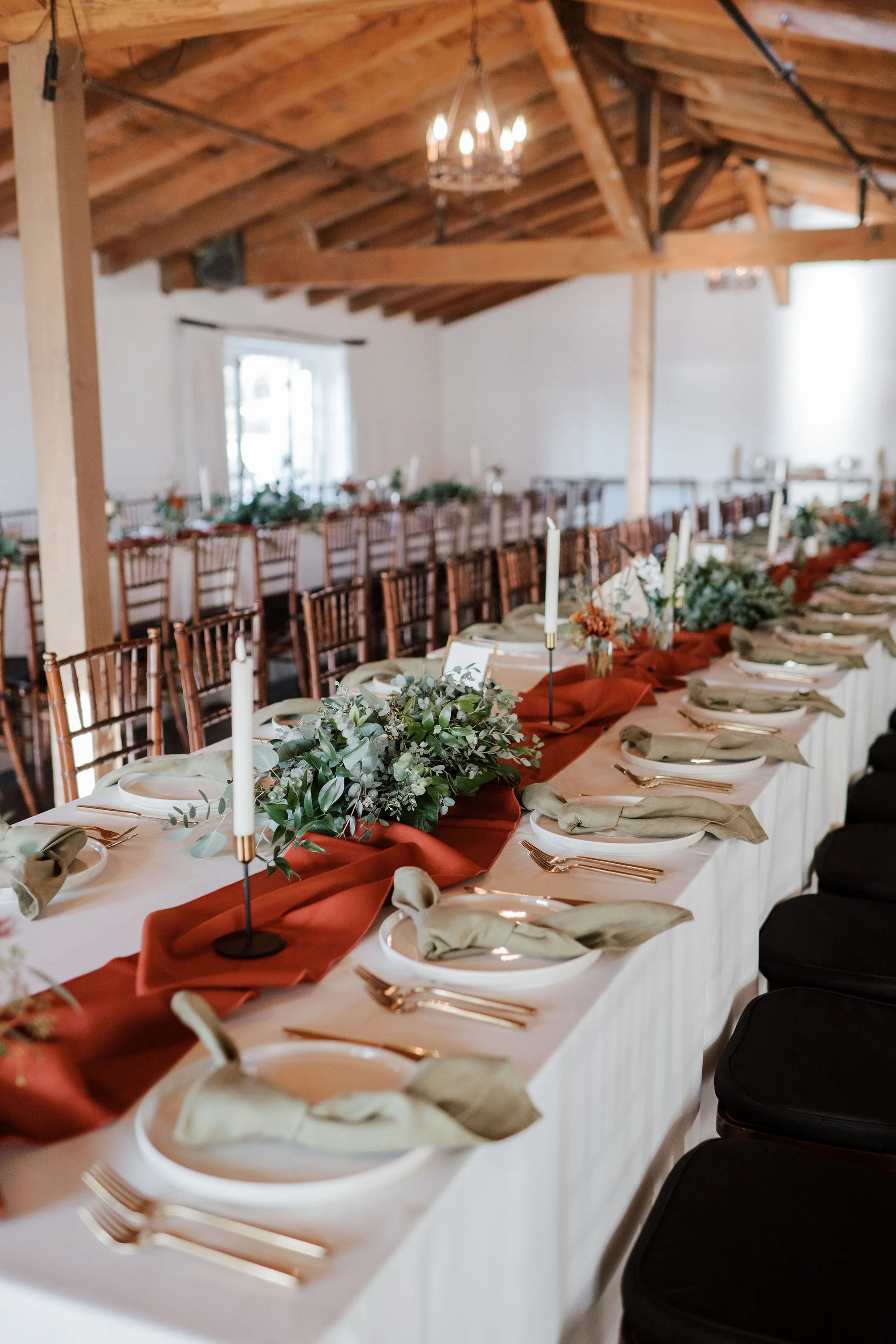 Elegant banquet table decorated with greenery, candles, and tableware in a rustic wooden venue.