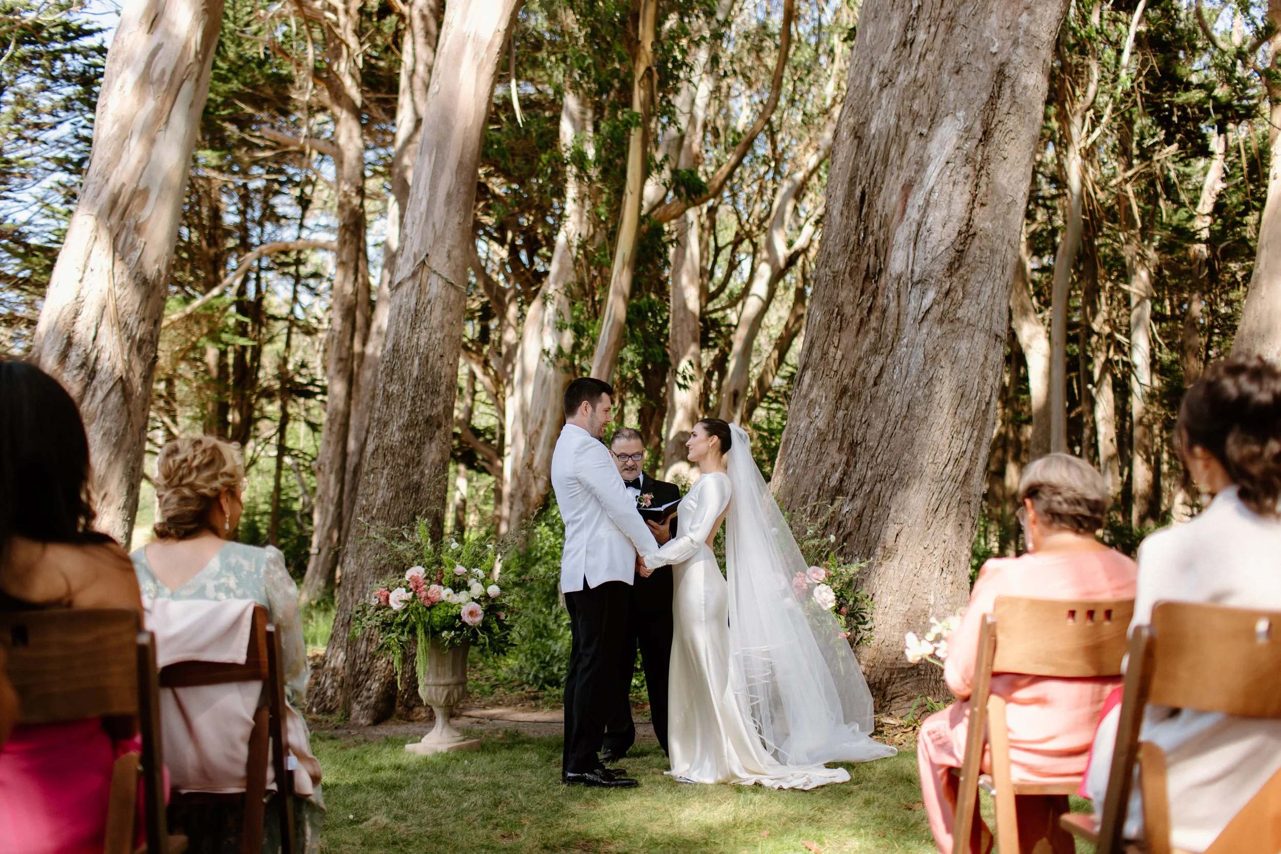 A wedding ceremony taking place outdoors among tall trees, with a bride and groom holding hands and facing each other, officiant in the middle, and guests seated on either side, all surrounded by nature.