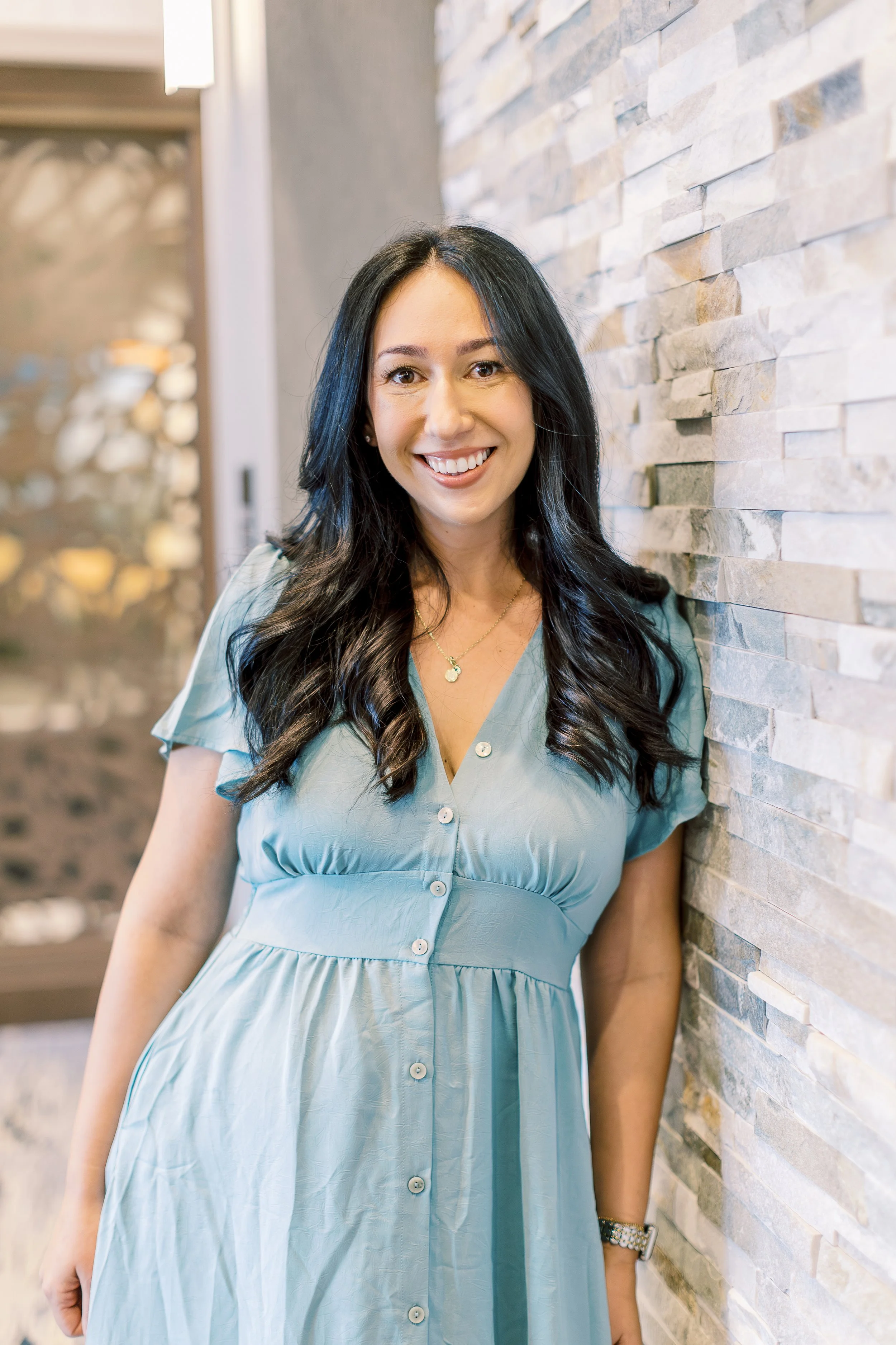 A woman with long black hair, smiling, wearing a light blue dress, standing against a stone wall in an indoor setting.