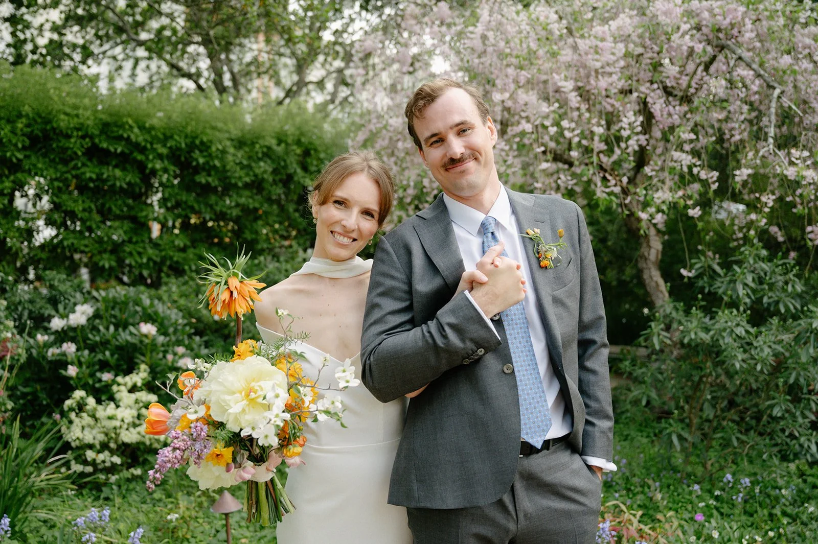 A newlywed couple smiling outdoors in a garden with blooming trees and various flowers, the woman holding a bouquet and the man dressed in a suit.