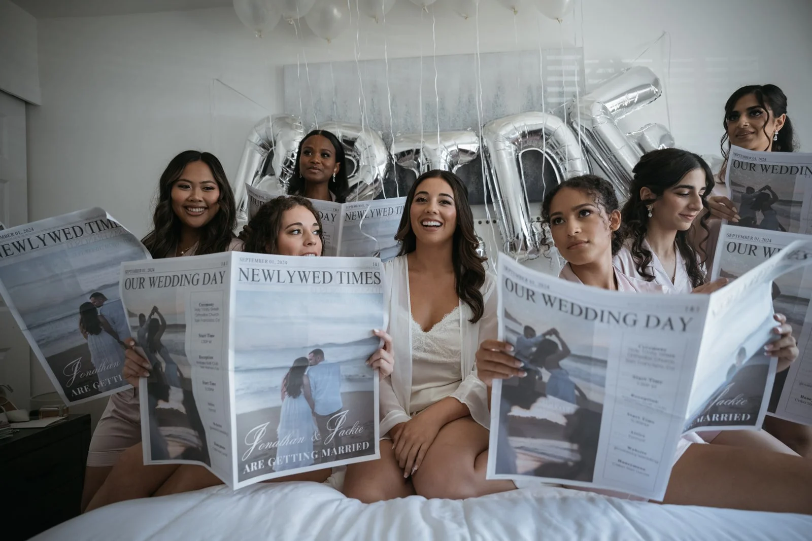 Group of women sitting on a bed reading newspapers about a wedding, with balloons spelling 'LOVE' in the background.