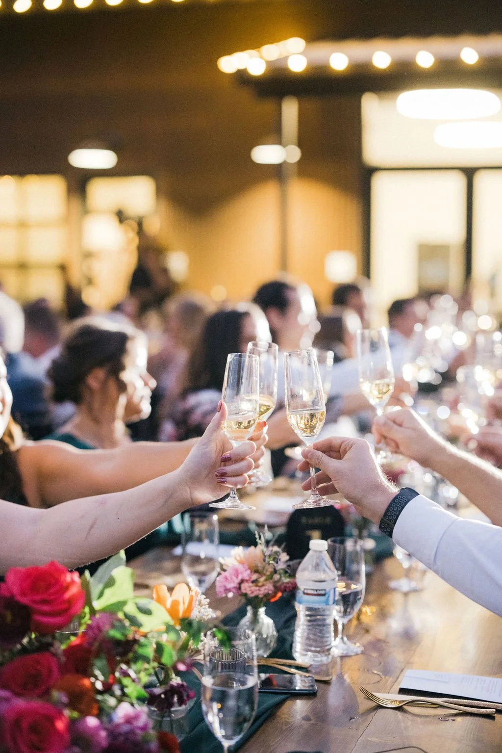 People celebrating at a wedding or banquet, toasting with glasses of white wine or champagne at a nicely decorated table with flowers.
