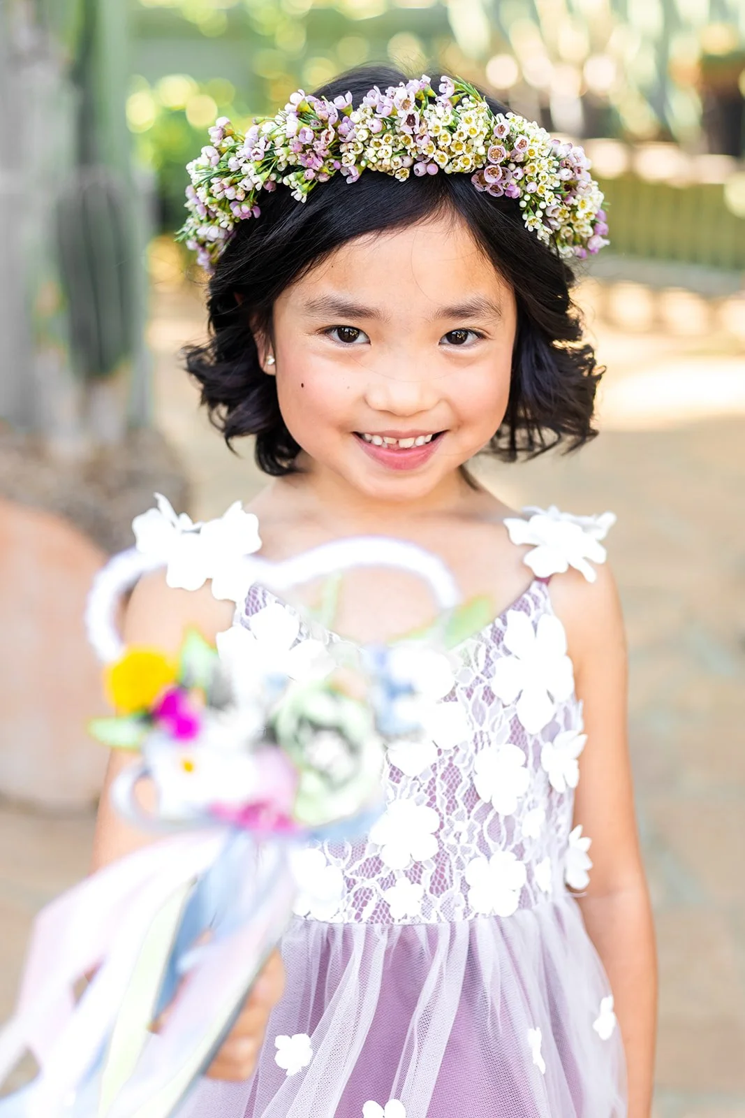 A young girl with dark hair wearing a purple floral princess dress and a flower crown, smiling outside and holding a wand.