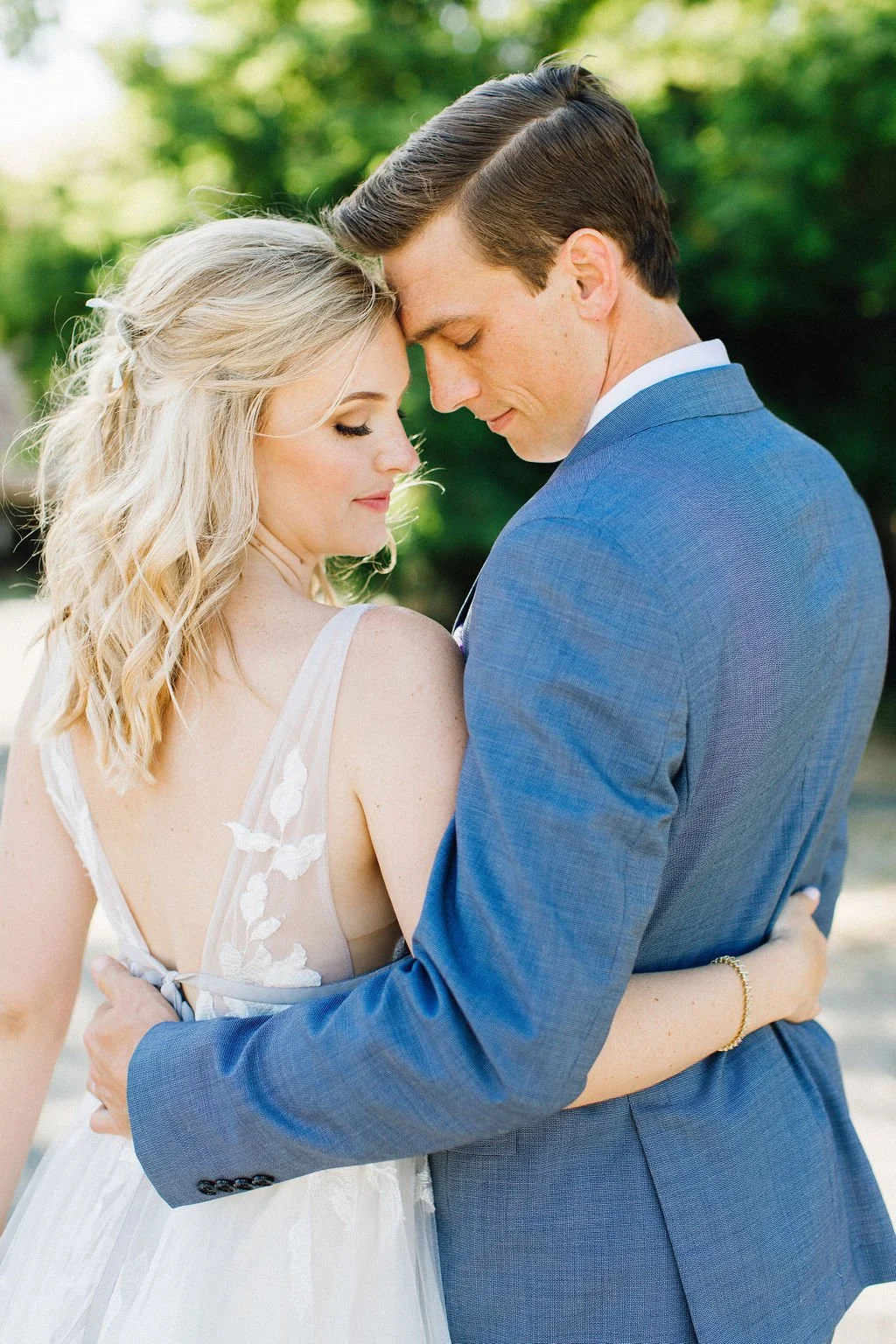 A bride and groom embrace outdoors, their foreheads touching and eyes closed, with trees and sunlight in the background.
