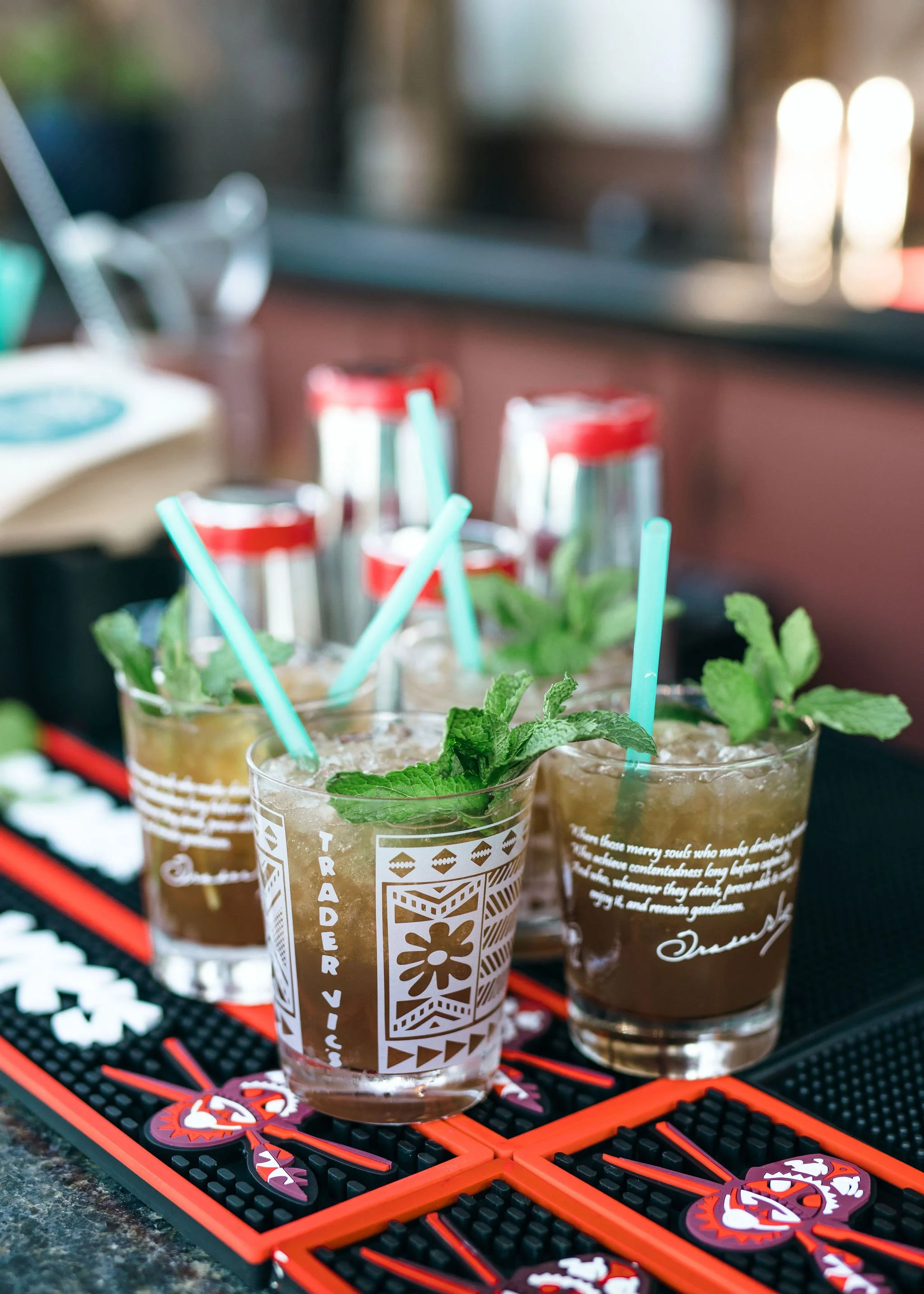 Three glasses of iced mojitos with mint leaves and blue straws on a black and orange bar mat, with Coca-Cola cans in the background.
