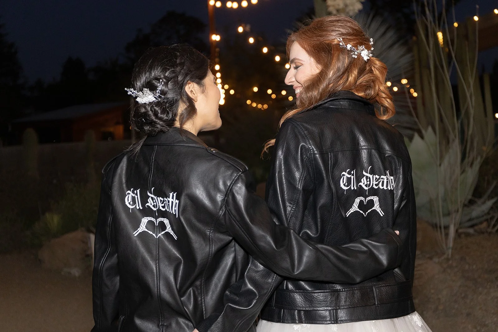 Two women wearing black leather jackets with 'El Death' written on the back, standing close and smiling at each other during the evening with string lights overhead.