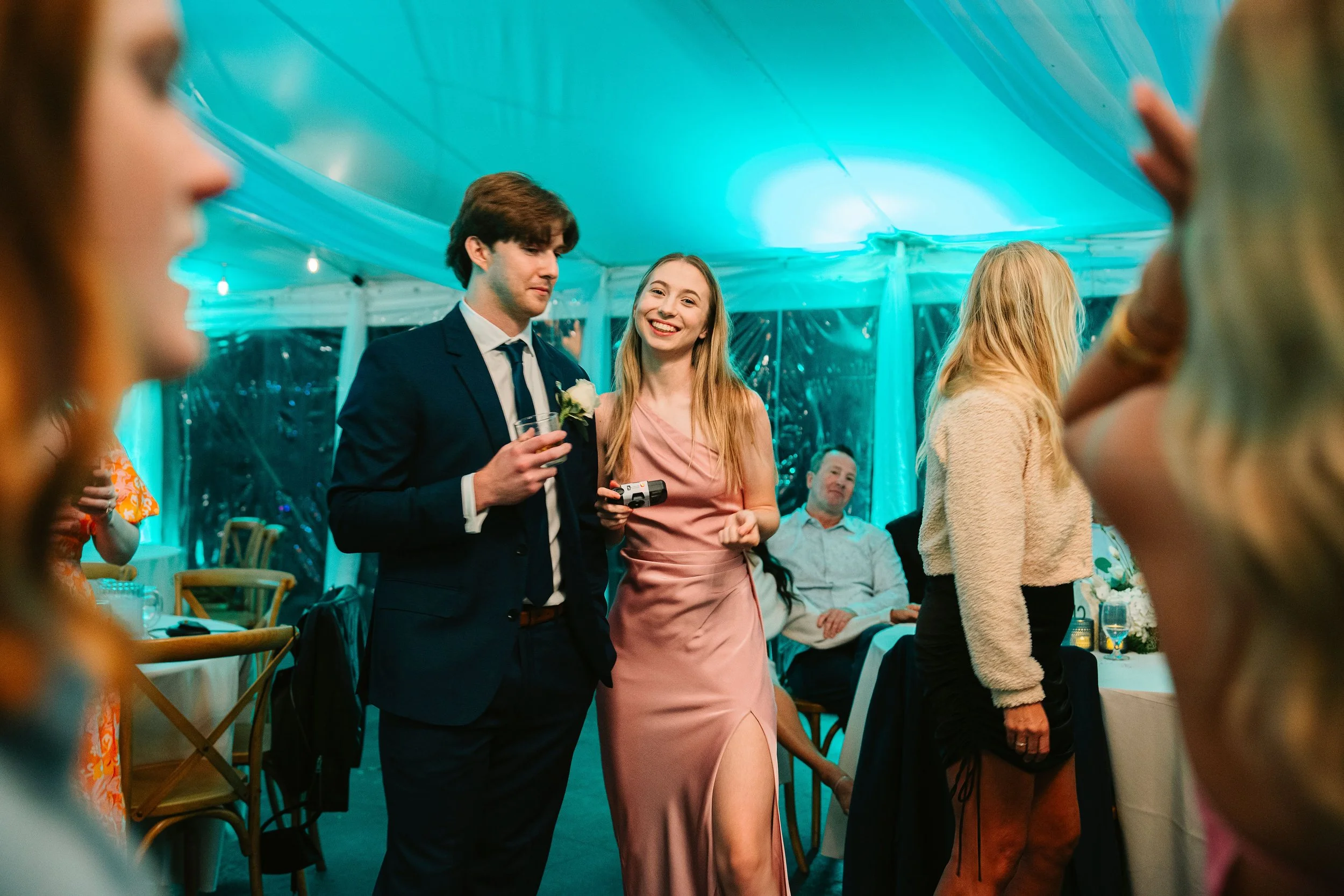 A group of people at a formal event or celebration in a decorated tent, with a man in a suit and a woman in a pink dress smiling and holding drinks, and other guests mingling around.