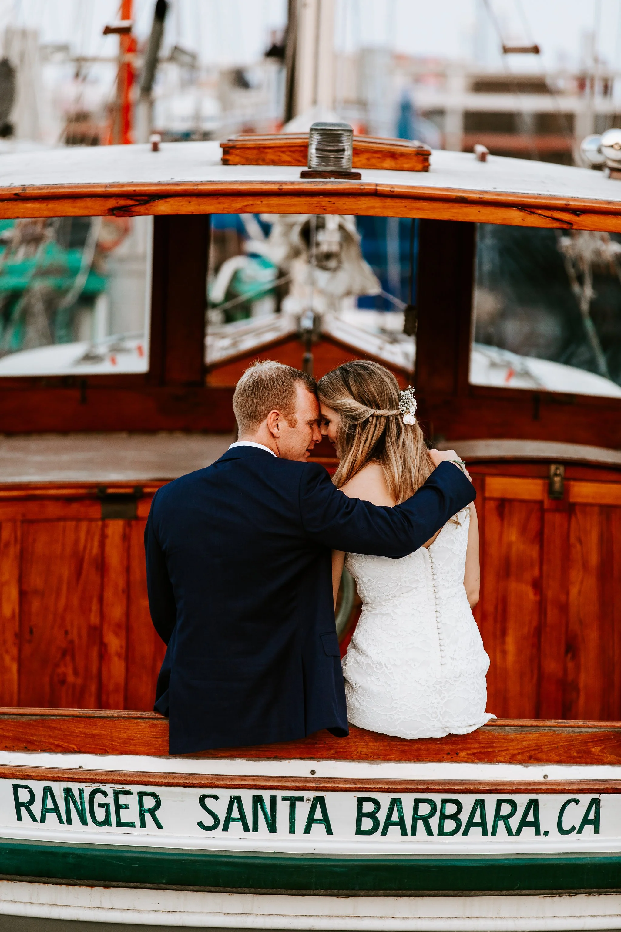 A couple in wedding attire sitting on the deck of a wooden boat named 'Ranger' in Santa Barbara, California, with their foreheads touching in an intimate moment.