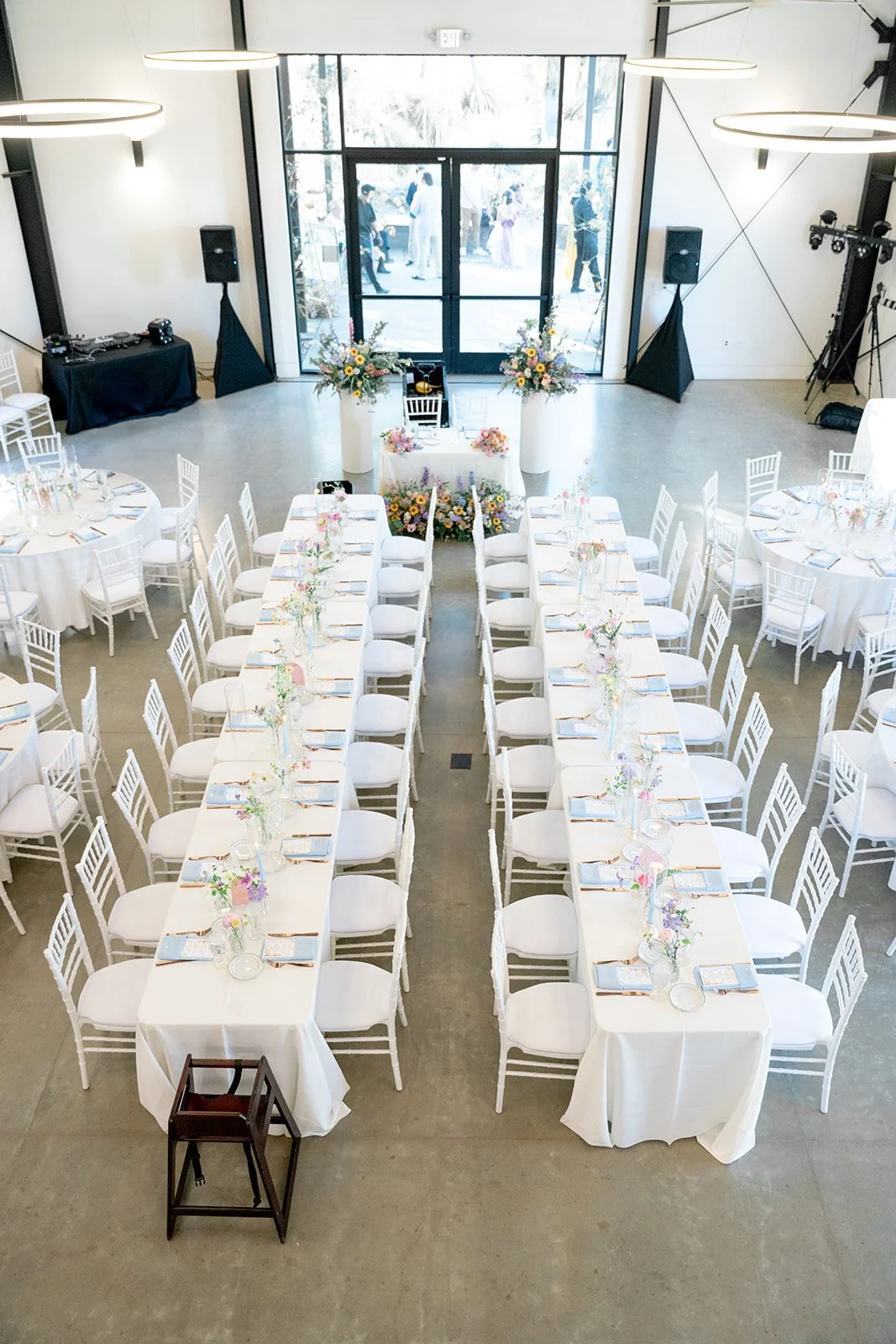 A beautifully decorated reception hall with long rectangular tables covered in white tablecloths, set with glassware, plates, and floral centerpieces. The hall has a large glass wall at the front, with guests visible outside, and is illuminated by modern circular hanging lights.