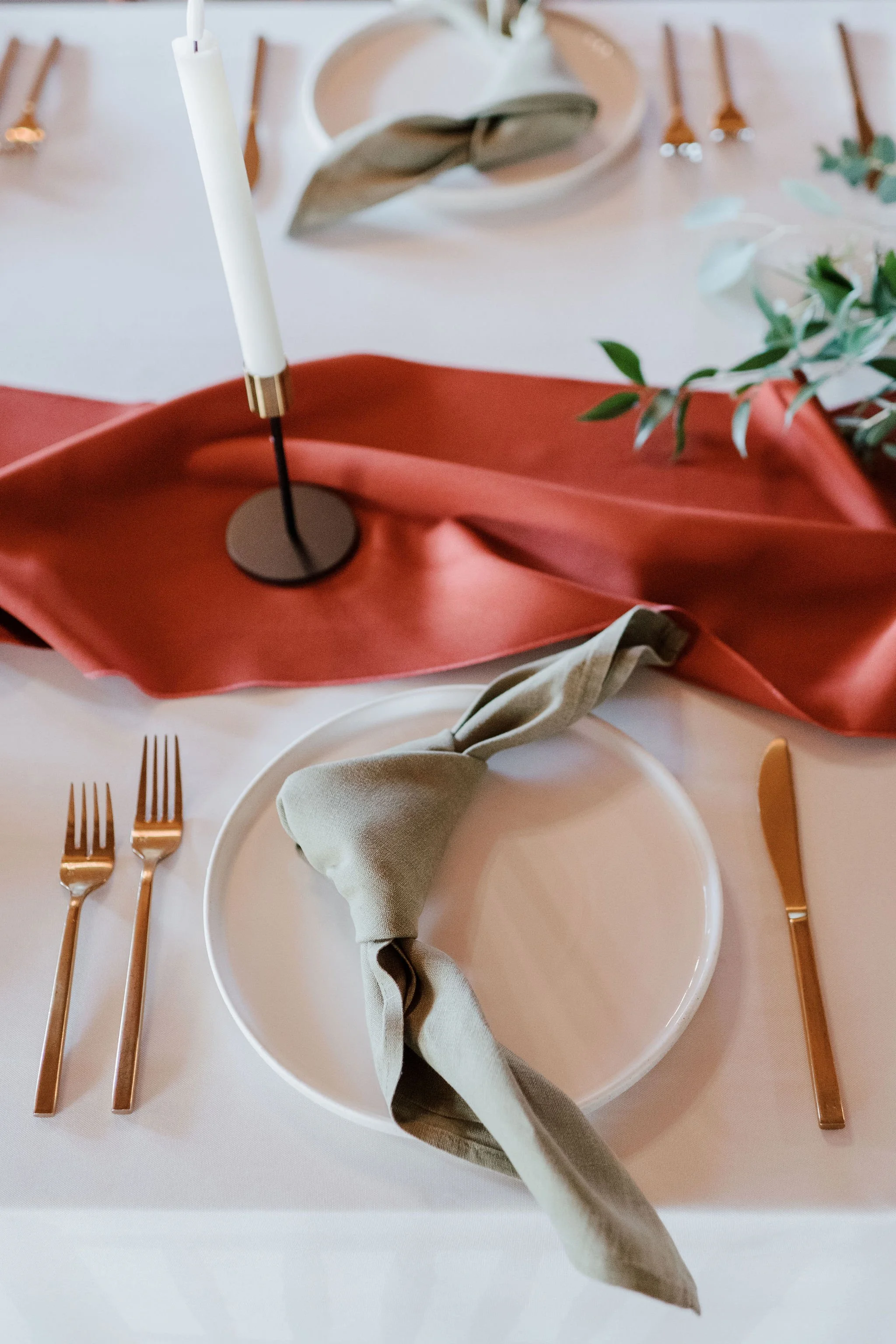 Elegant table setting with white plate, green cloth napkin tied around, rose gold cutlery, a black candle holder with a white candle, a red satin table runner, and decorative greenery.