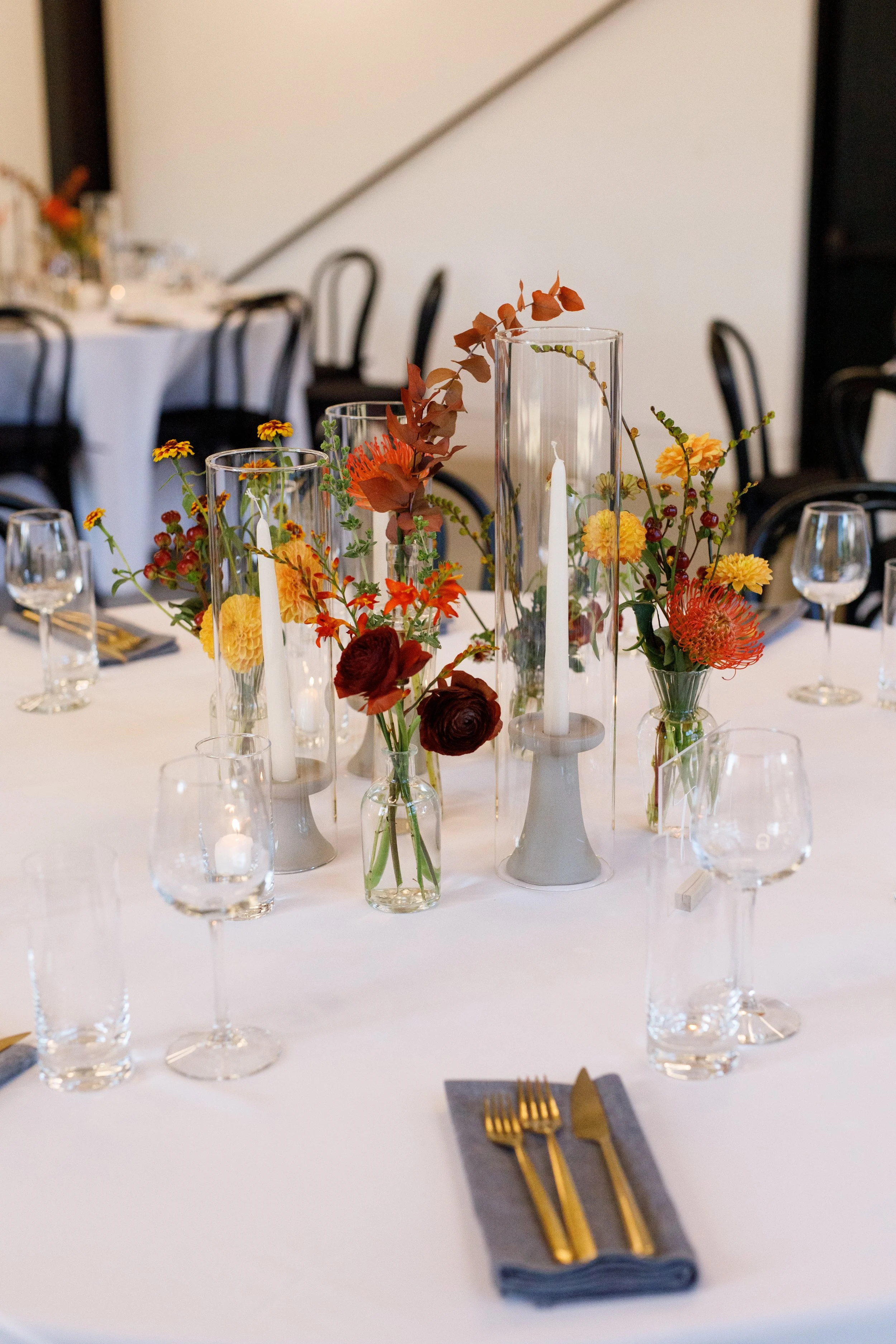 Elegant table centerpiece featuring tall glass cylinders with white candles and orange, red, and yellow flowers, set on a white tablecloth with glassware and gold cutlery.