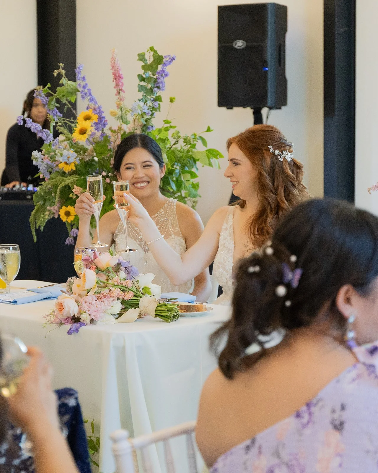 Two women at a wedding celebration toasting with champagne glasses, surrounded by floral arrangements and a DJ in the background.