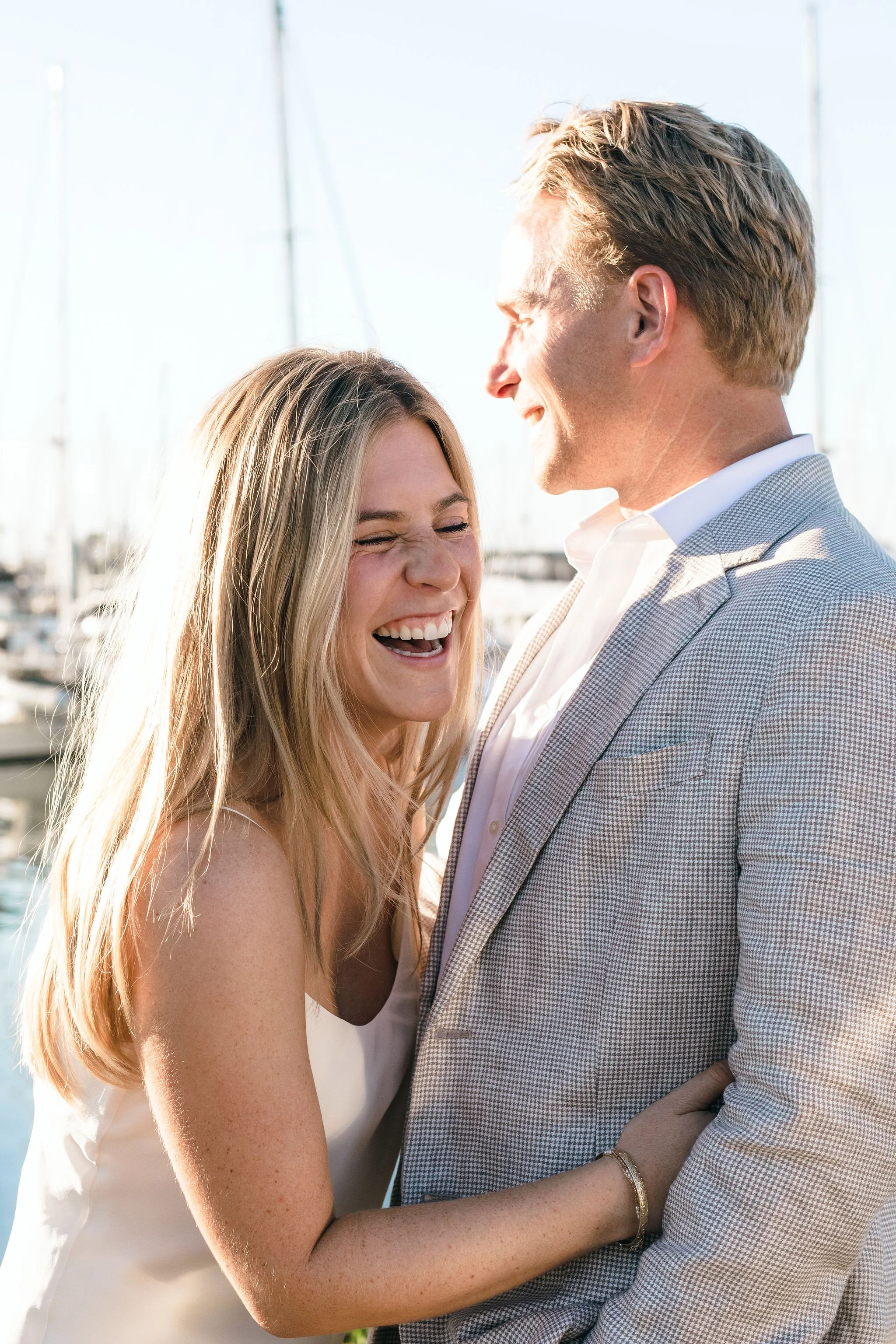 A joyful couple laughing together near a marina with sailboats in the background, sunny weather.