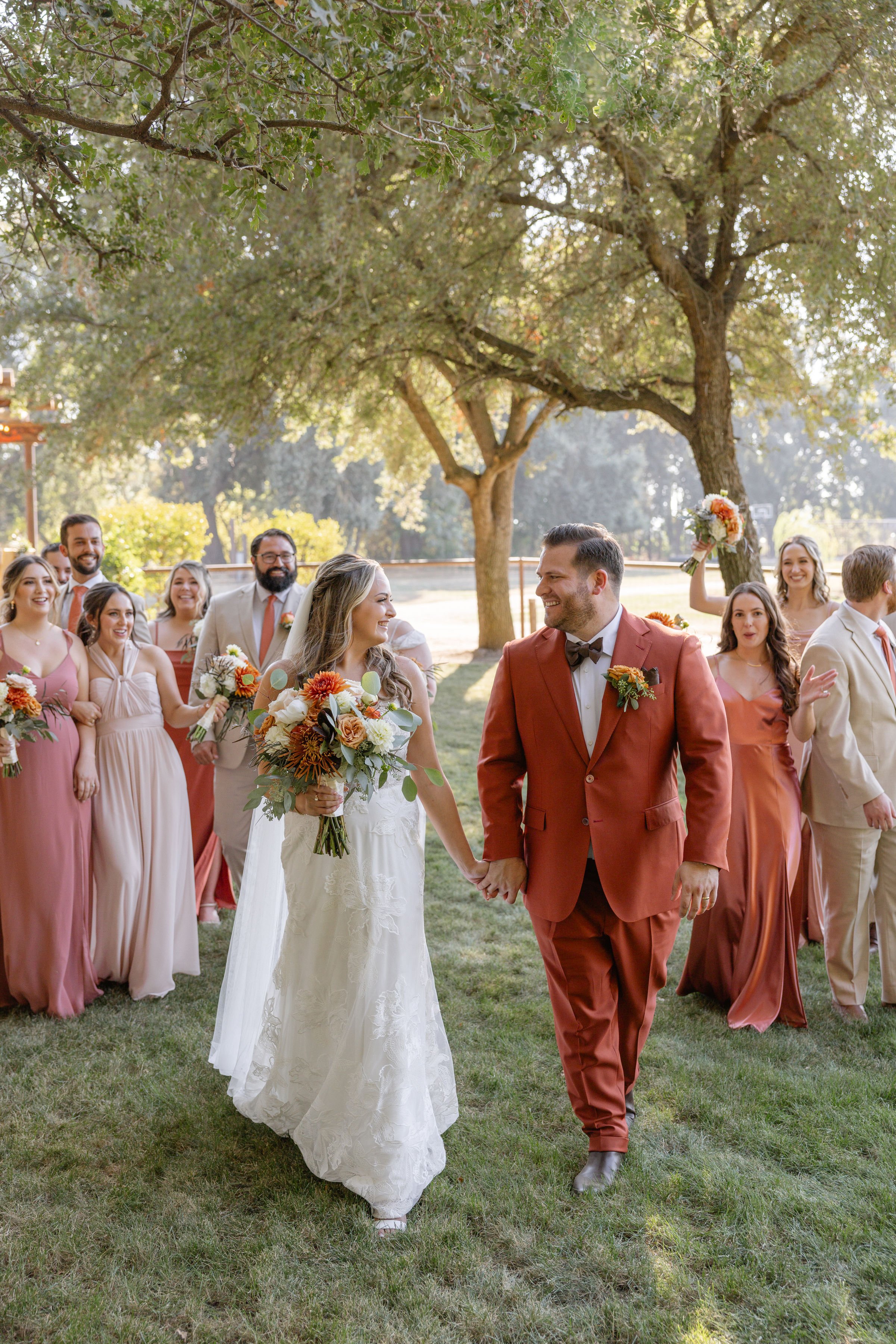 Bride and groom walking hand in hand outdoors, wedding party celebrating behind them, with large trees and sunlight.