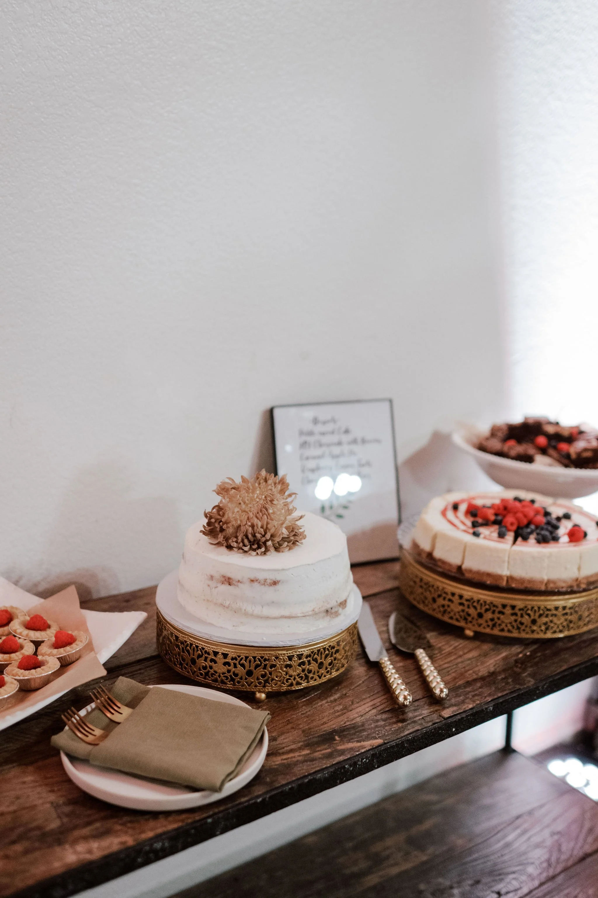 Dessert table with a white frosted cake decorated with a flower on top, cheesecake with fruit topping, and other desserts, set on a rustic wooden table with a beige wall background.