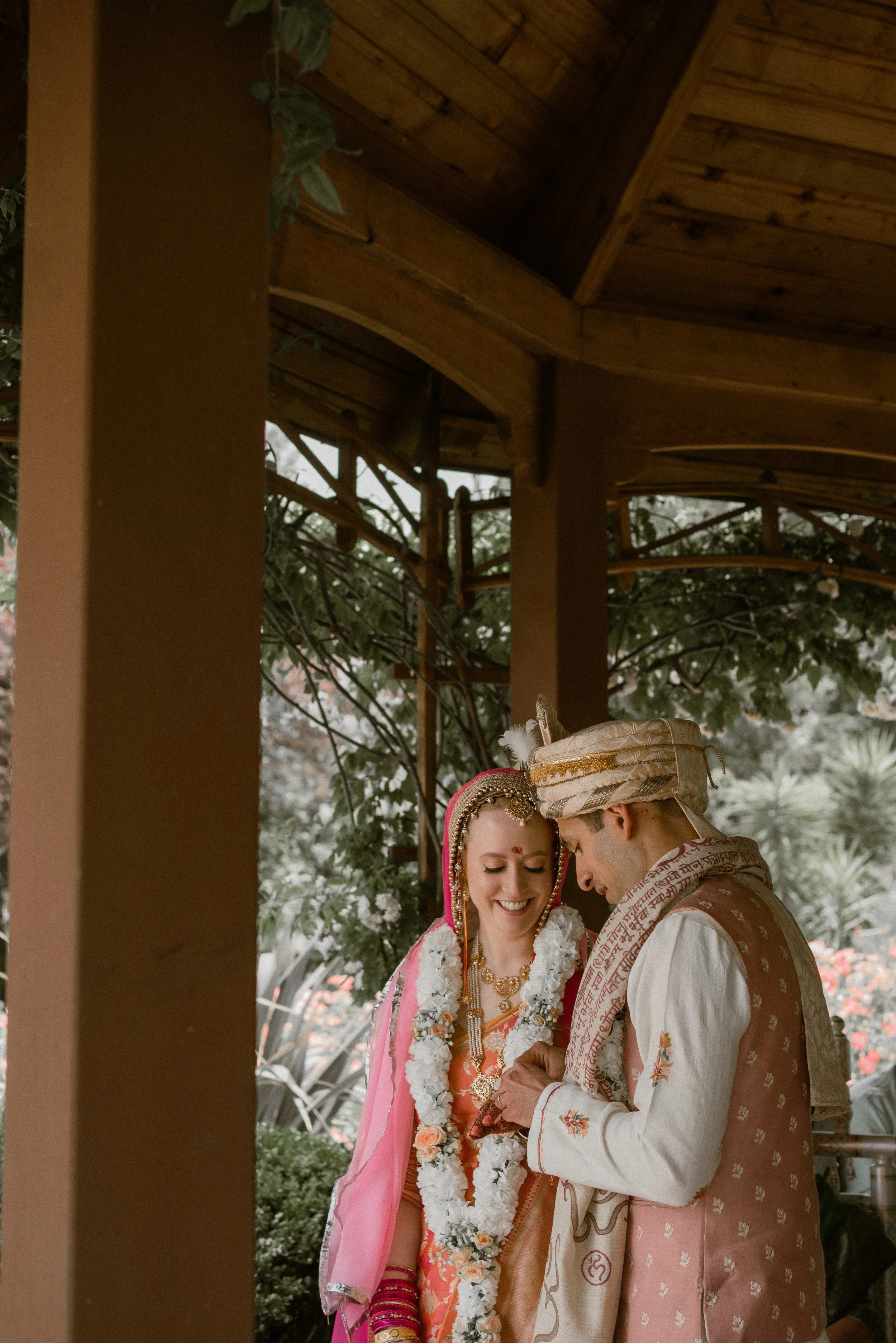 A bride and groom in traditional Indian wedding attire, standing together under a wooden gazebo, smiling and looking at a phone.