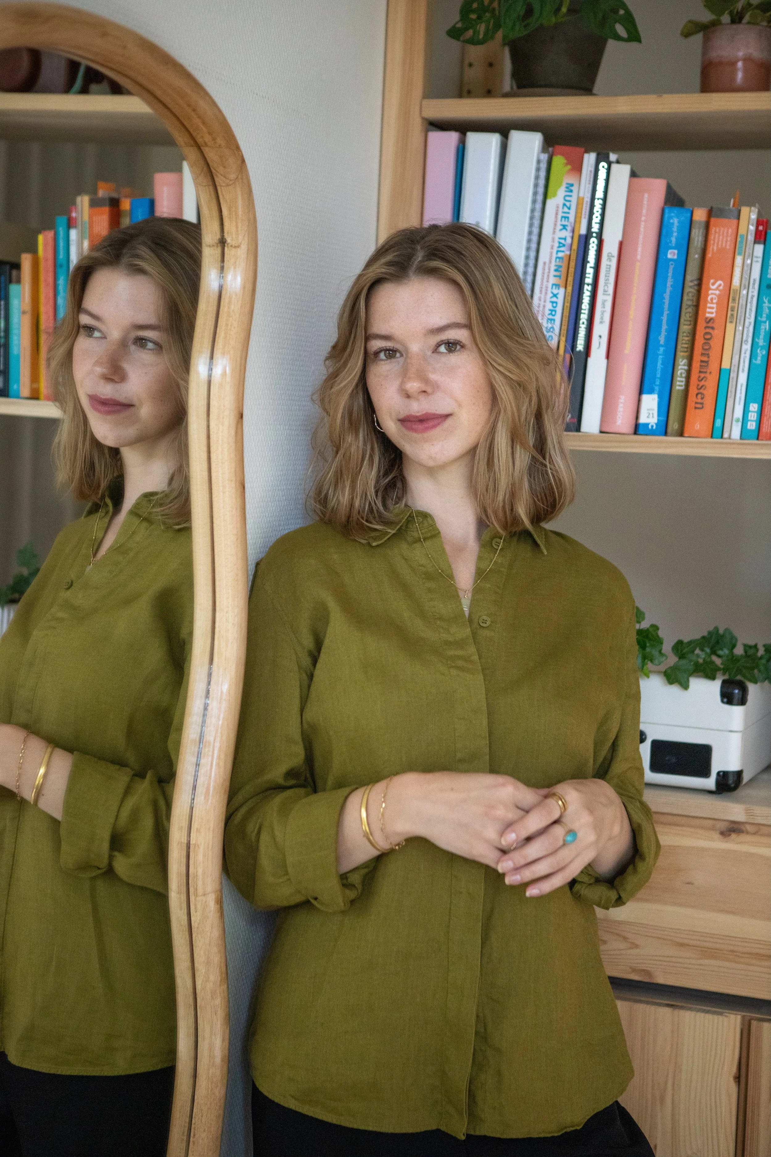 Een vrouw met schouderlange, krullende, lichtbruin haar, gekleed in een groen shirt, staat naast een houten kast met boeken en planten, en kijkt in de spiegel.