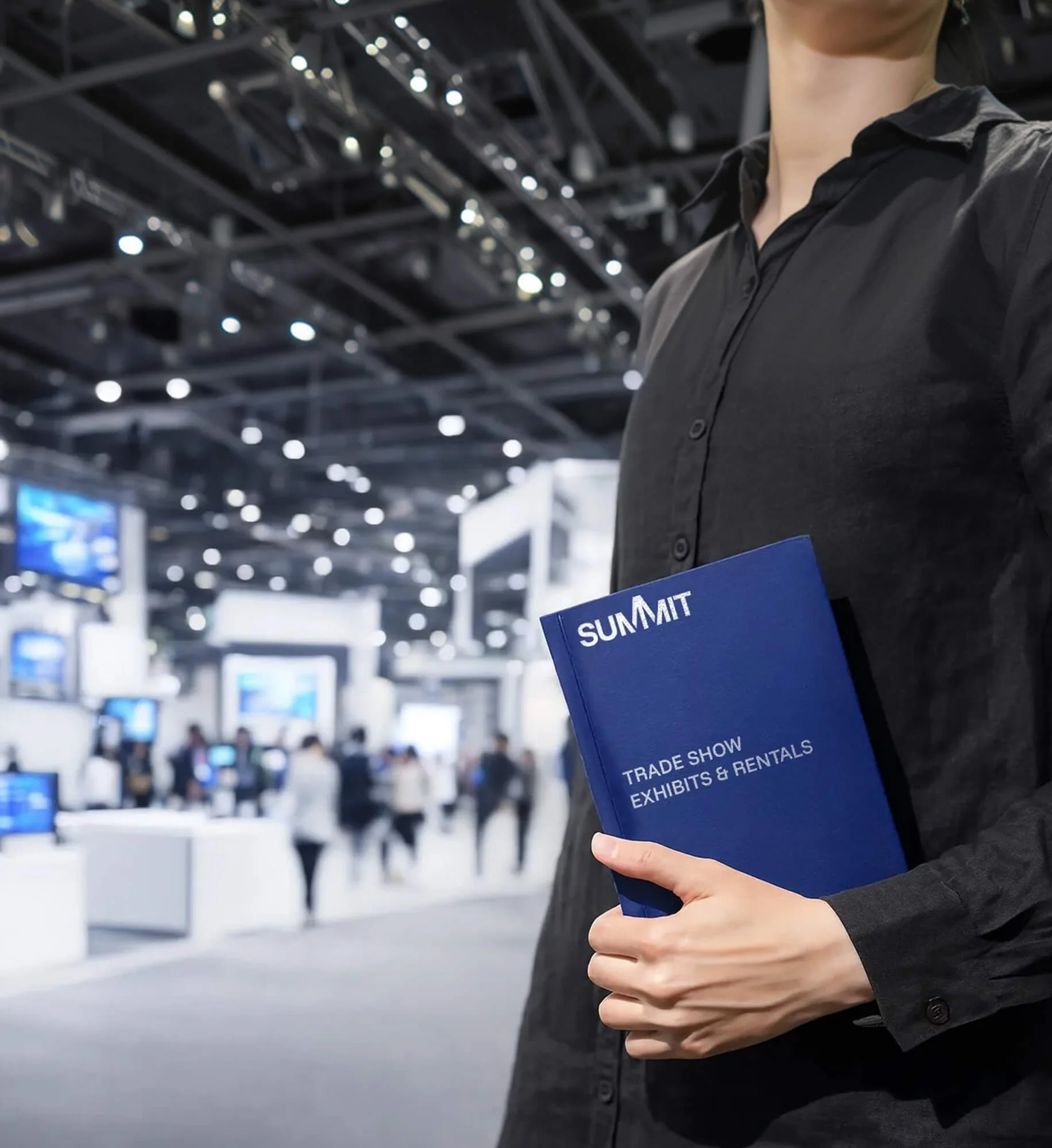 Person holding a blue brochure titled 'Summit: Trade Show Exhibits & Rentals' at a trade show event. The background shows a busy exhibition hall with people, displays, and bright overhead lighting.
