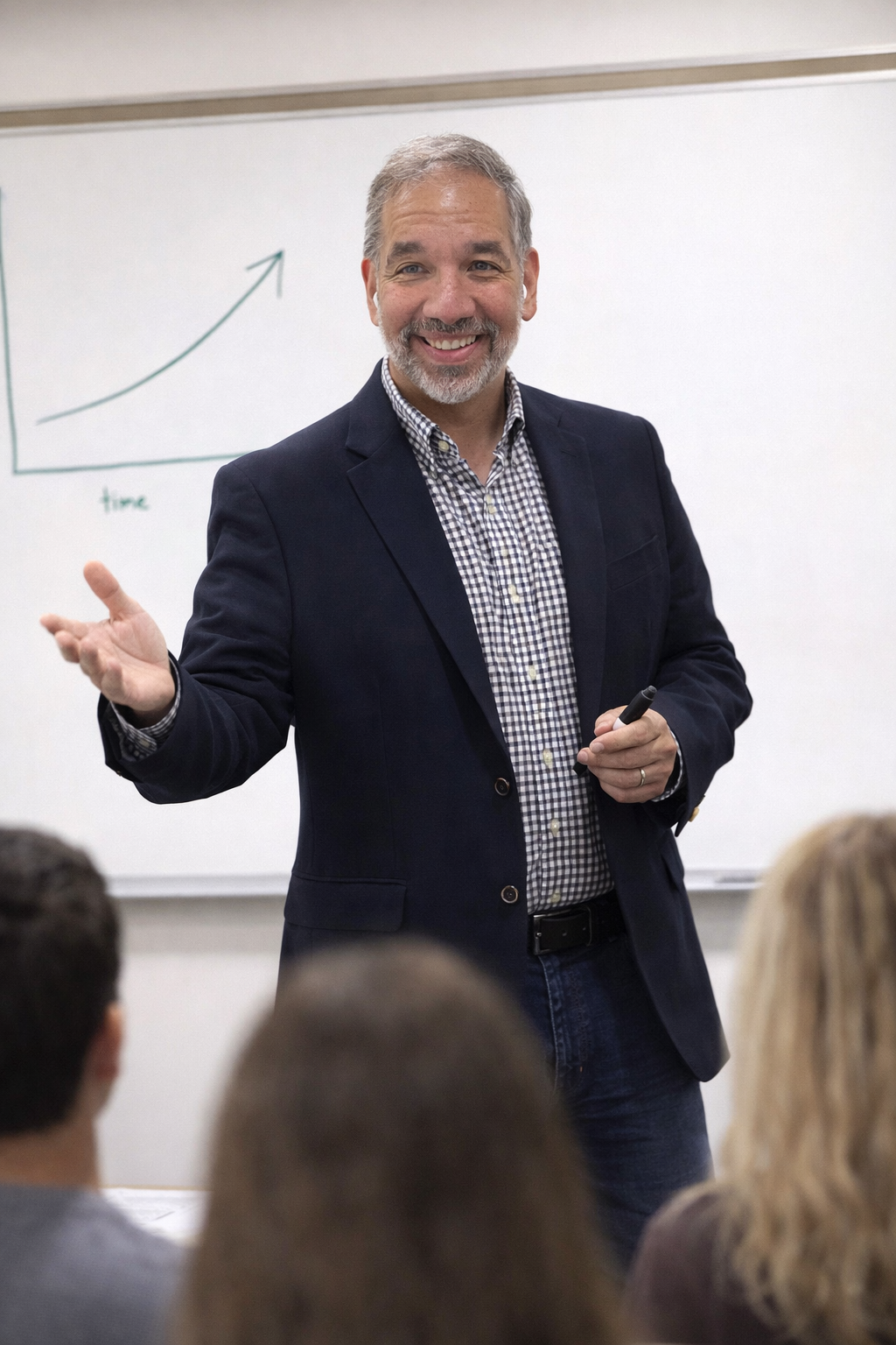 A middle-aged man with gray hair and beard, dressed in a checkered shirt and dark blazer, smiling and gesturing with his left hand while holding a marker in his right hand in front of a whiteboard with a graph showing an upward trend labeled 'time'.