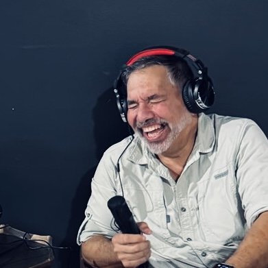 A man with gray hair and a beard, wearing headphones and a light-colored shirt, singing or speaking into a microphone with a joyful expression.