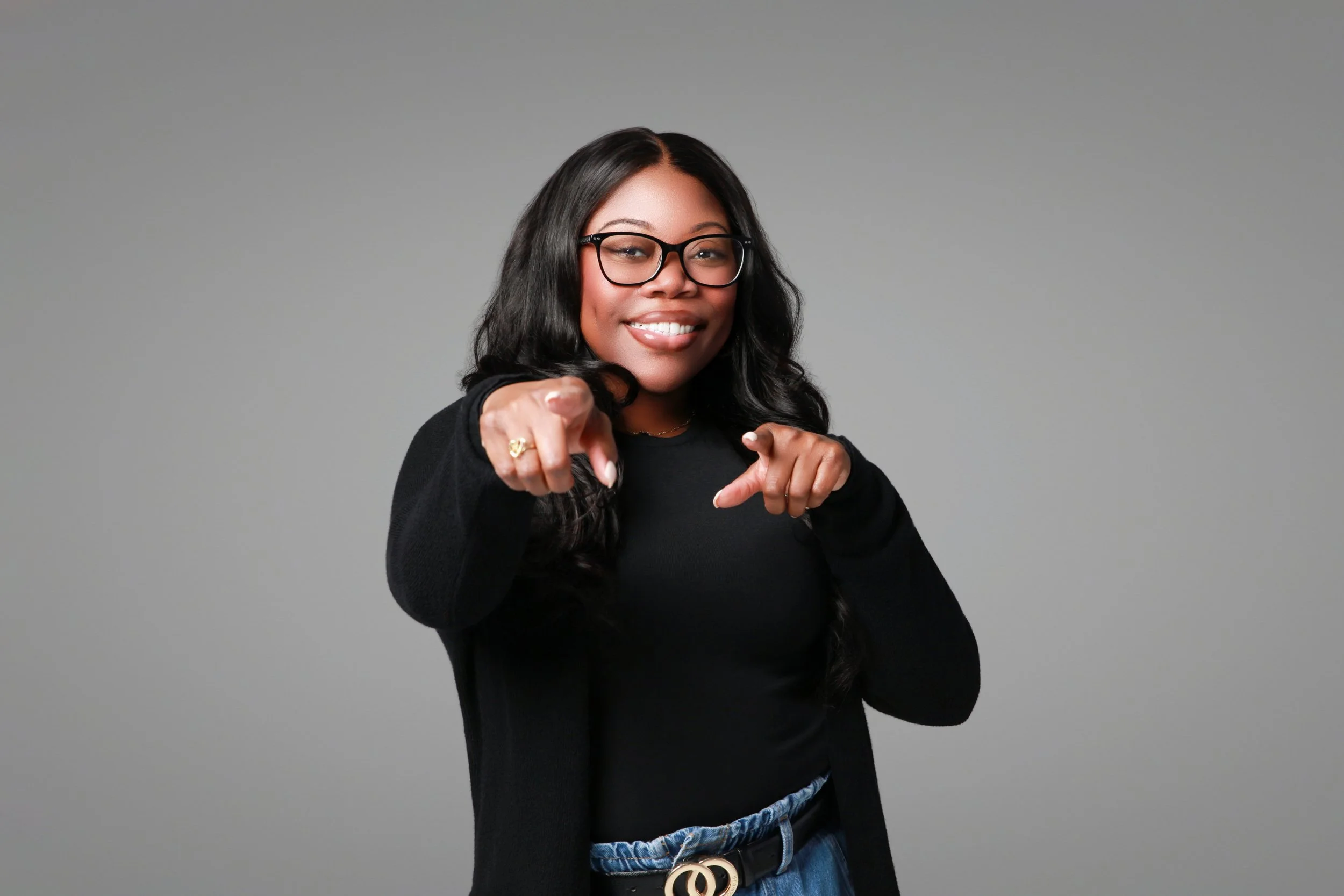 A woman with long black hair, glasses, and a black top, smiling and pointing at the camera against a plain gray background.