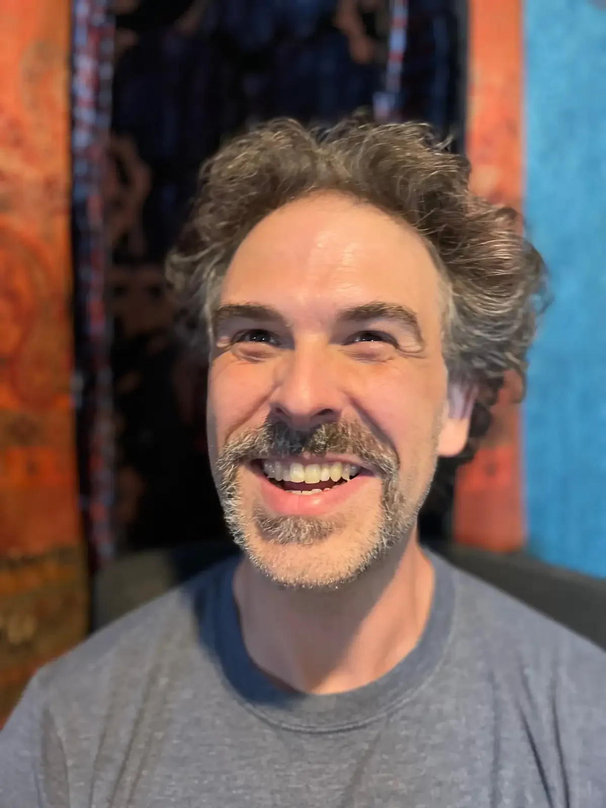 A smiling man with curly gray hair and a beard, wearing a gray t-shirt, stands in front of colorful textured artwork.