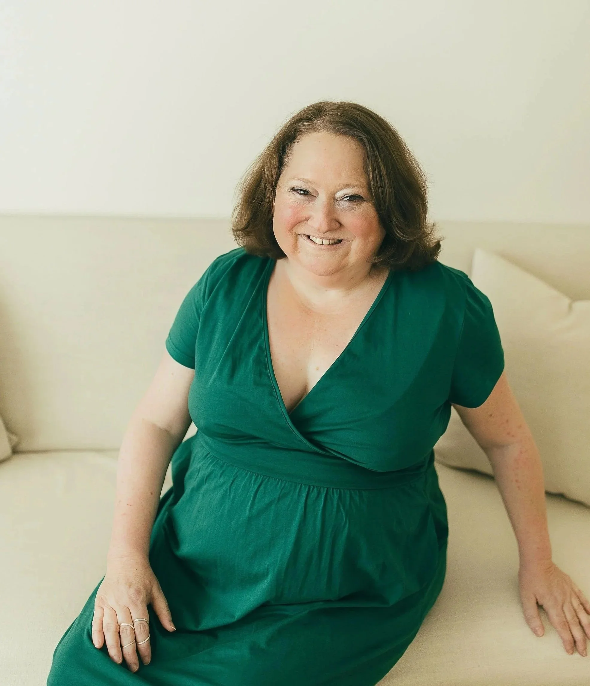 A woman with shoulder-length brown hair, smiling, wearing a green dress, sitting on a beige surface, with a white wall in the background.