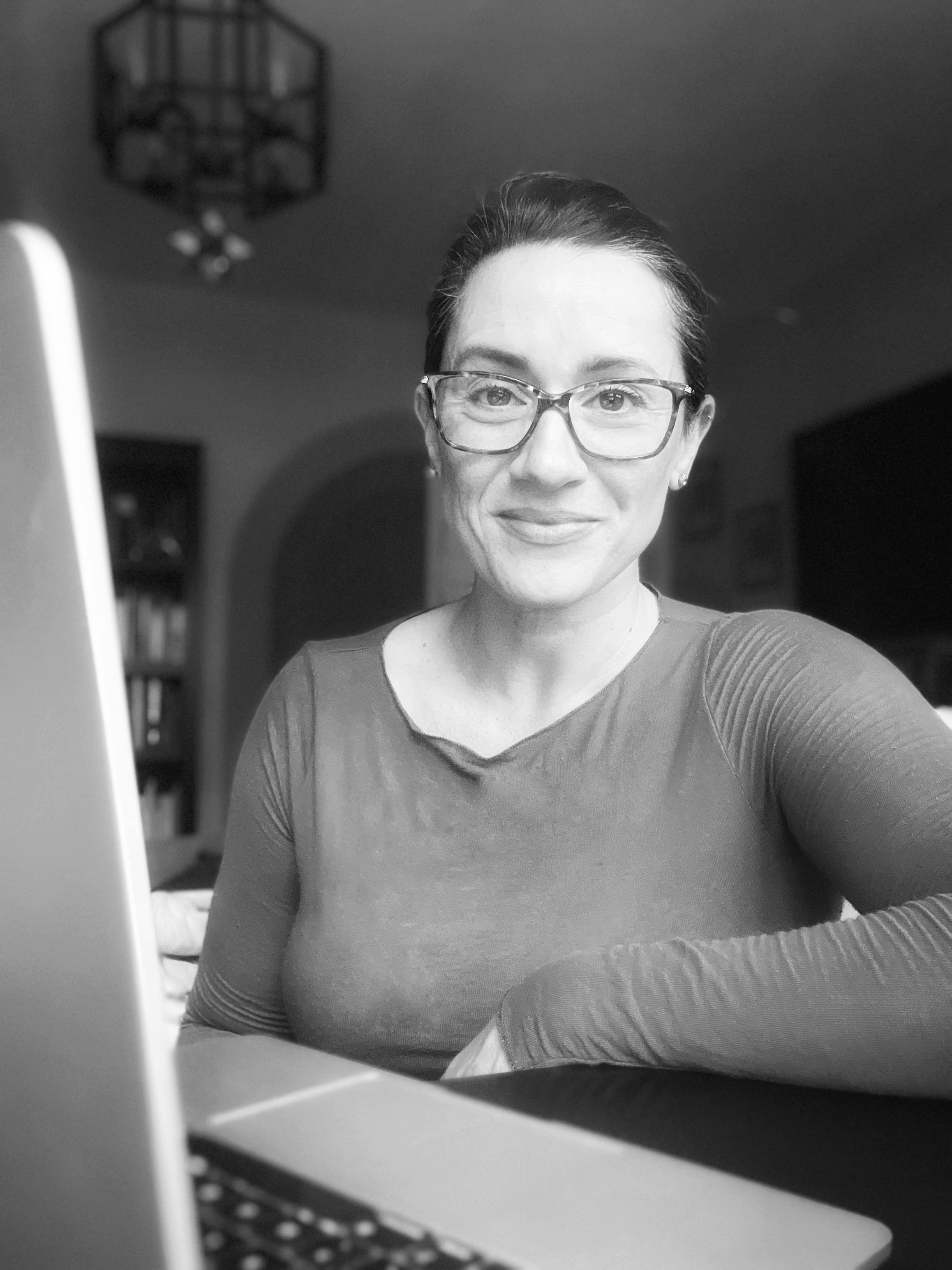 A woman with glasses smiling at the camera, sitting near a laptop in a home environment.