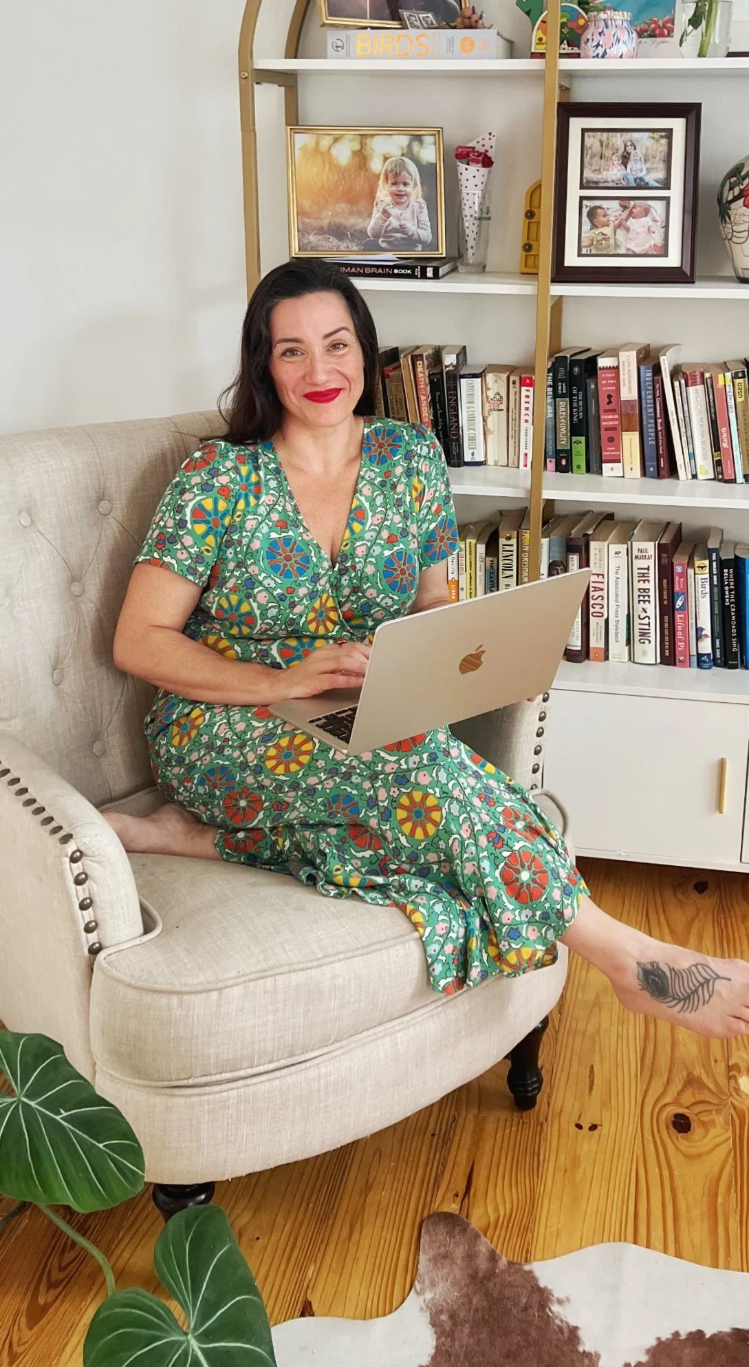 A woman with dark hair and red lipstick sits on a beige armchair, working on a silver MacBook. She wears a colorful, patterned dress and has a tattoo on her left ankle. Behind her is a white bookshelf filled with books, framed photographs, and decorative items. A large green plant is visible in the foreground.