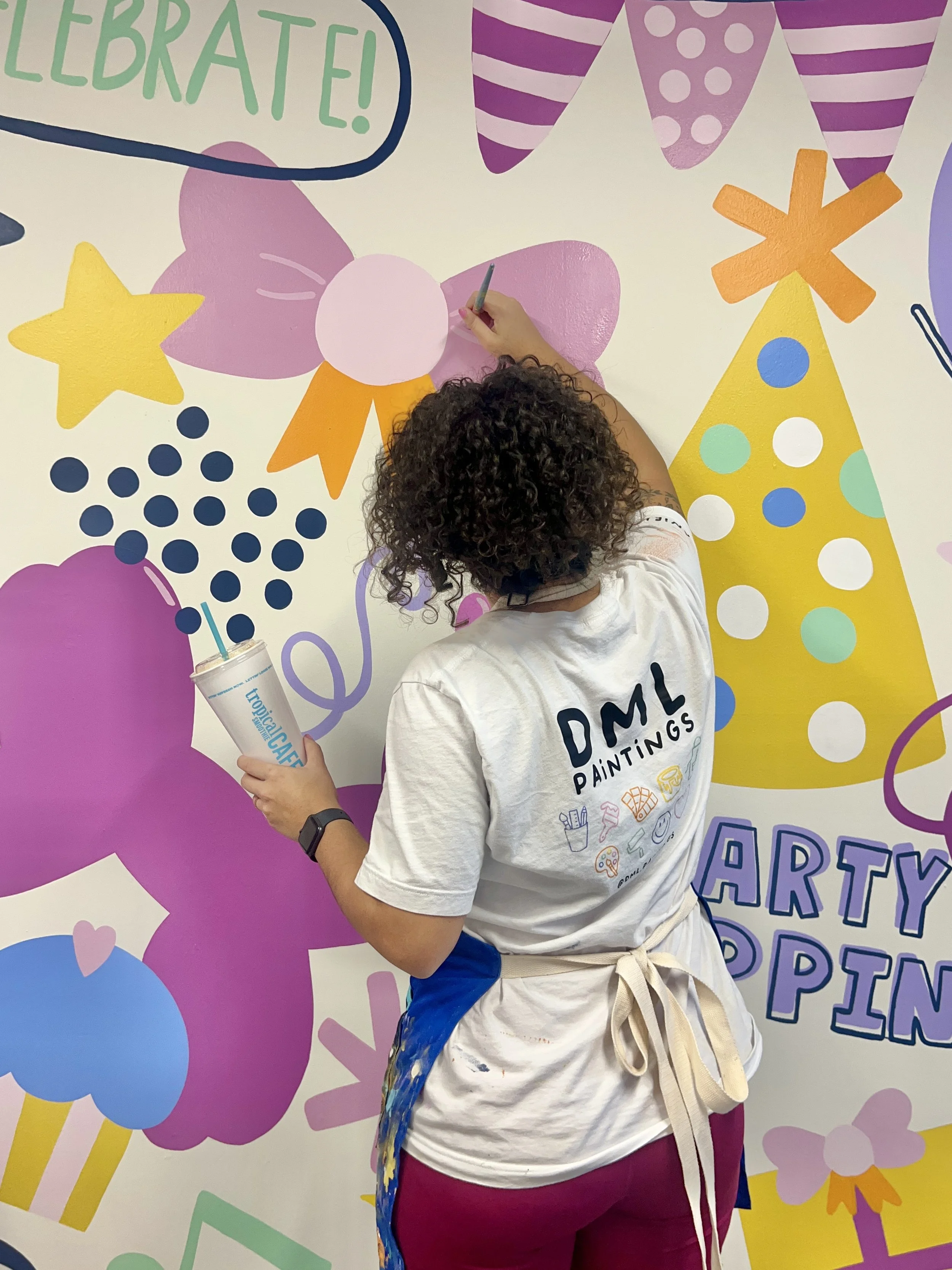 Person painting a pink flower on a colorful party-themed wall with balloons, stars, and banners.