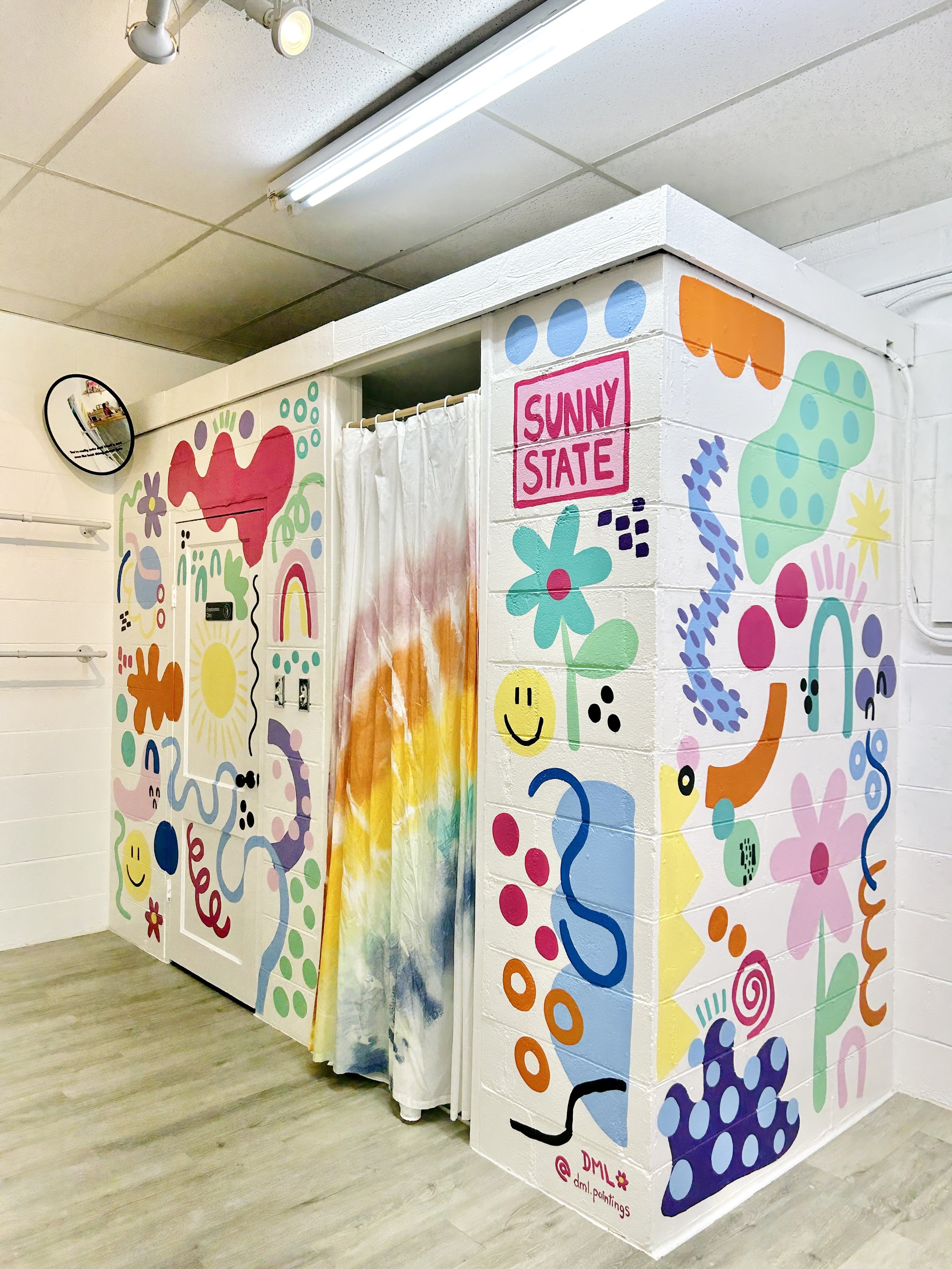 Colorful mural on the walls depicting abstract shapes, flowers, smiley faces, and playful designs surrounding a small dressing room with a rainbow-colored curtain. The top right corner has a pink sign that reads "Sunny State."
