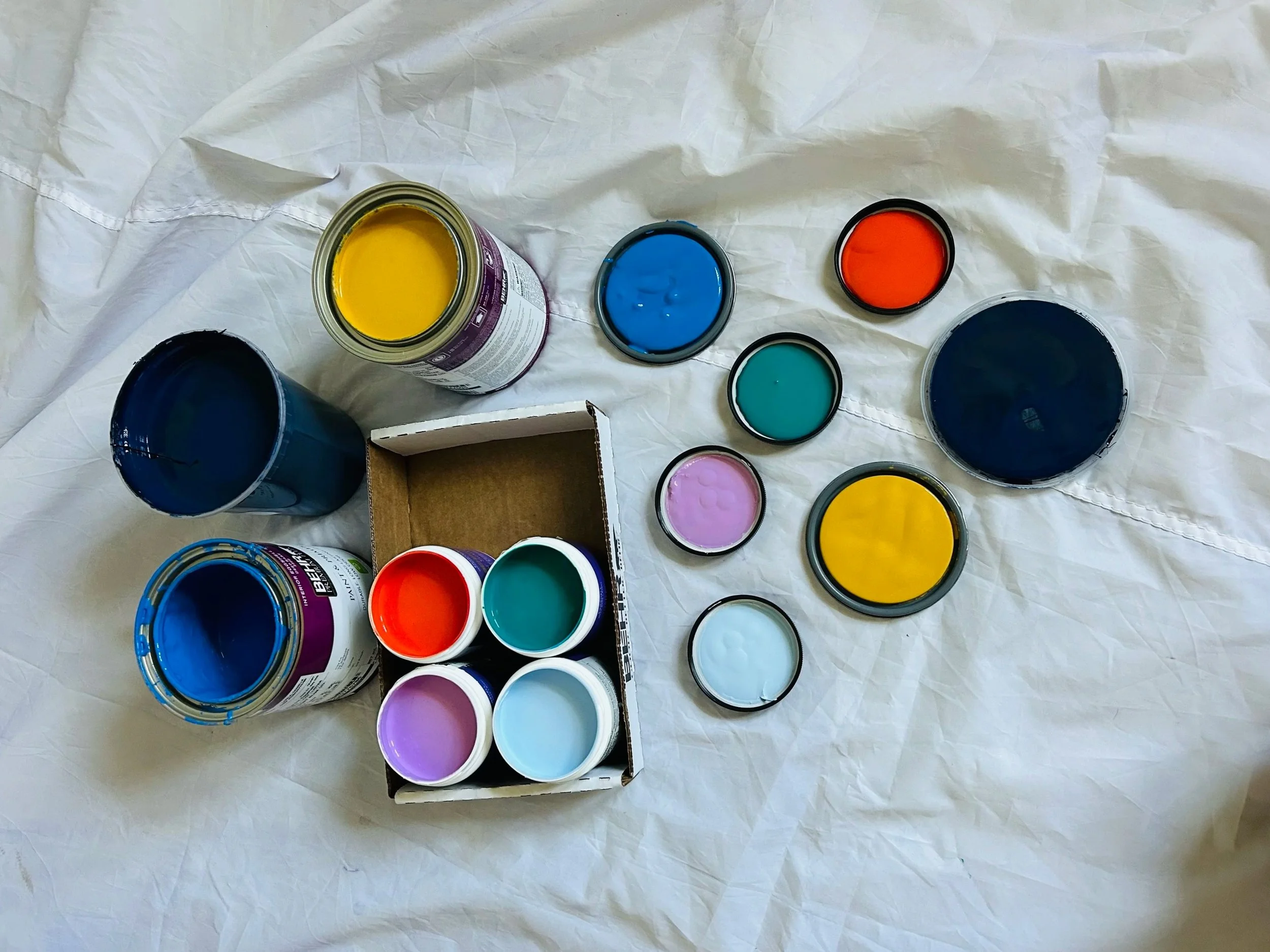 Open cans of colored paint in yellow, blue, teal, purple, white, and red, with some cans on a paper surface and a cardboard box holding four cans of paint.