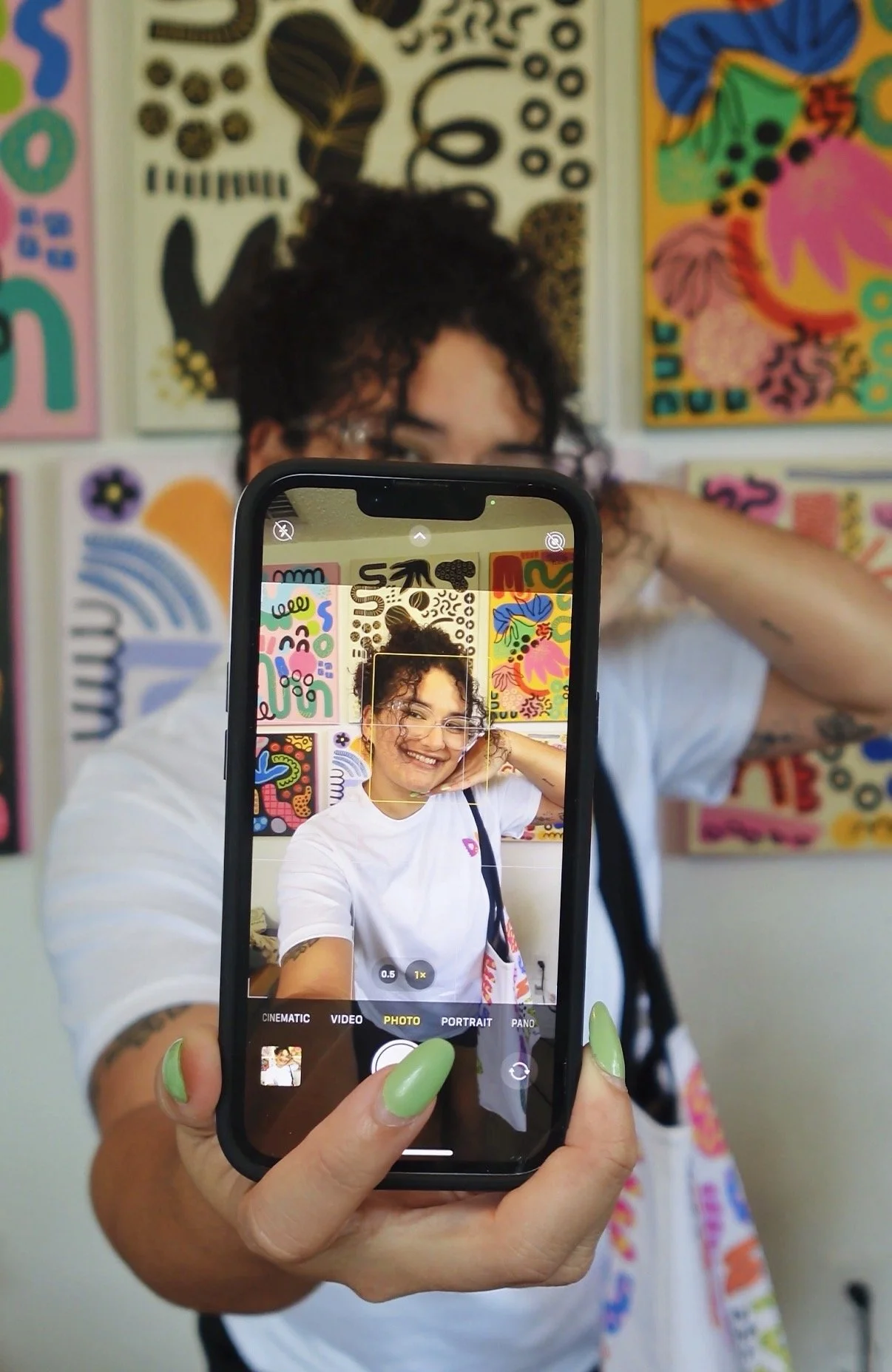 A woman holding a smartphone takes a selfie in front of colorful abstract artwork on the wall.