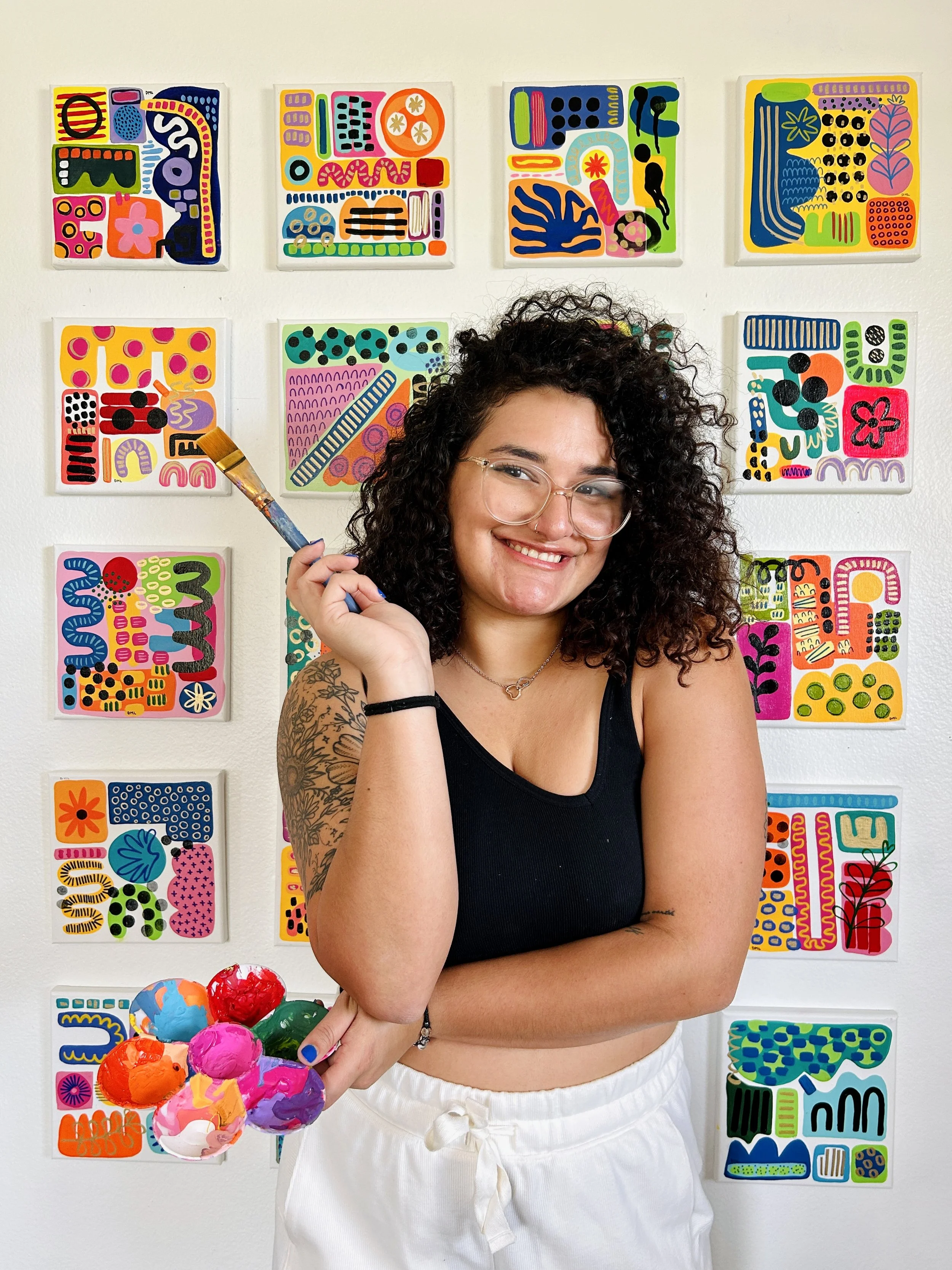 Young woman with curly hair and glasses holding a paintbrush and palette, standing in front of colorful abstract art on gallery wall.