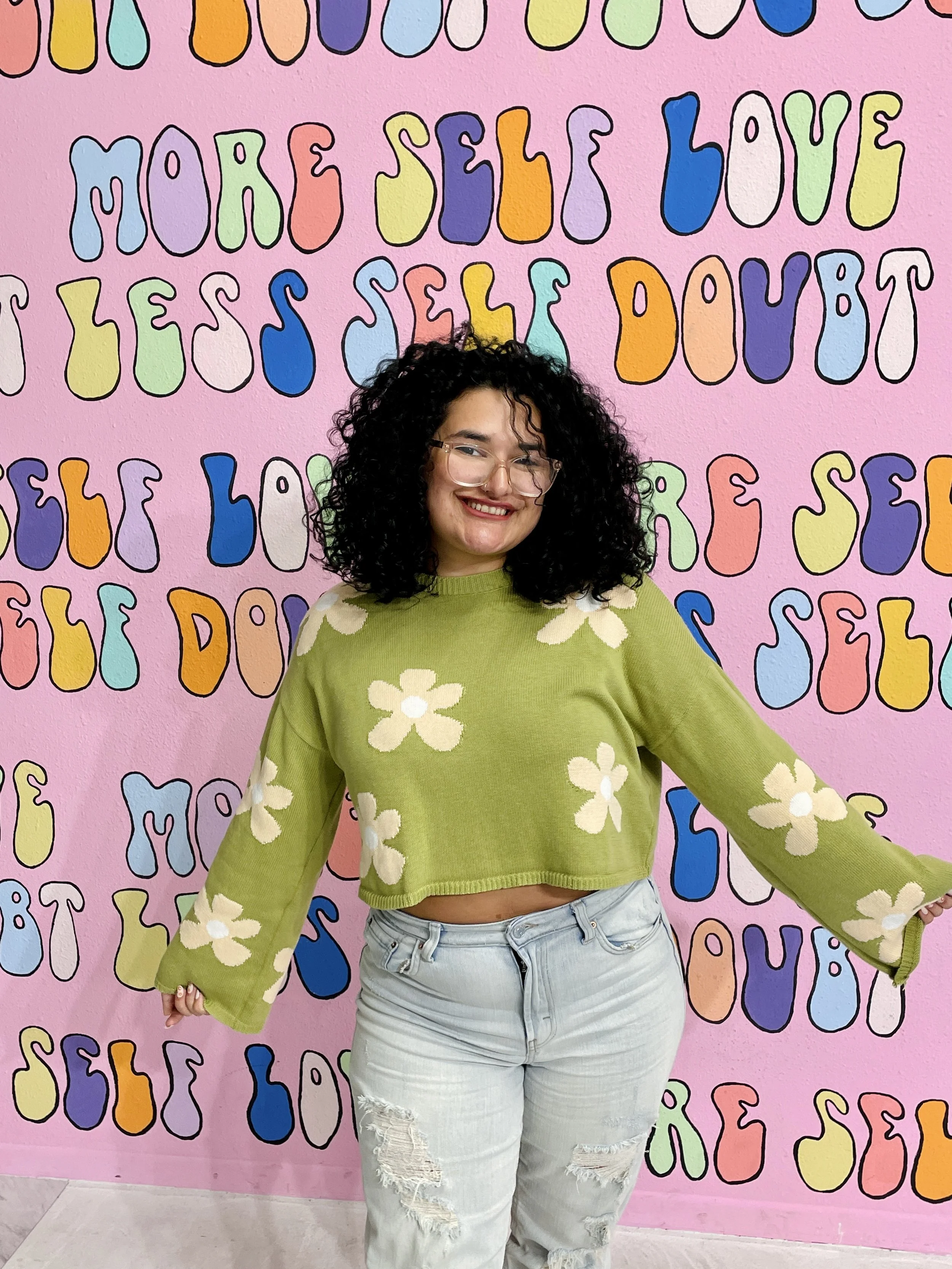 A young woman with curly black hair and glasses, smiling and posing in front of a pink wall with colorful, artistic text.