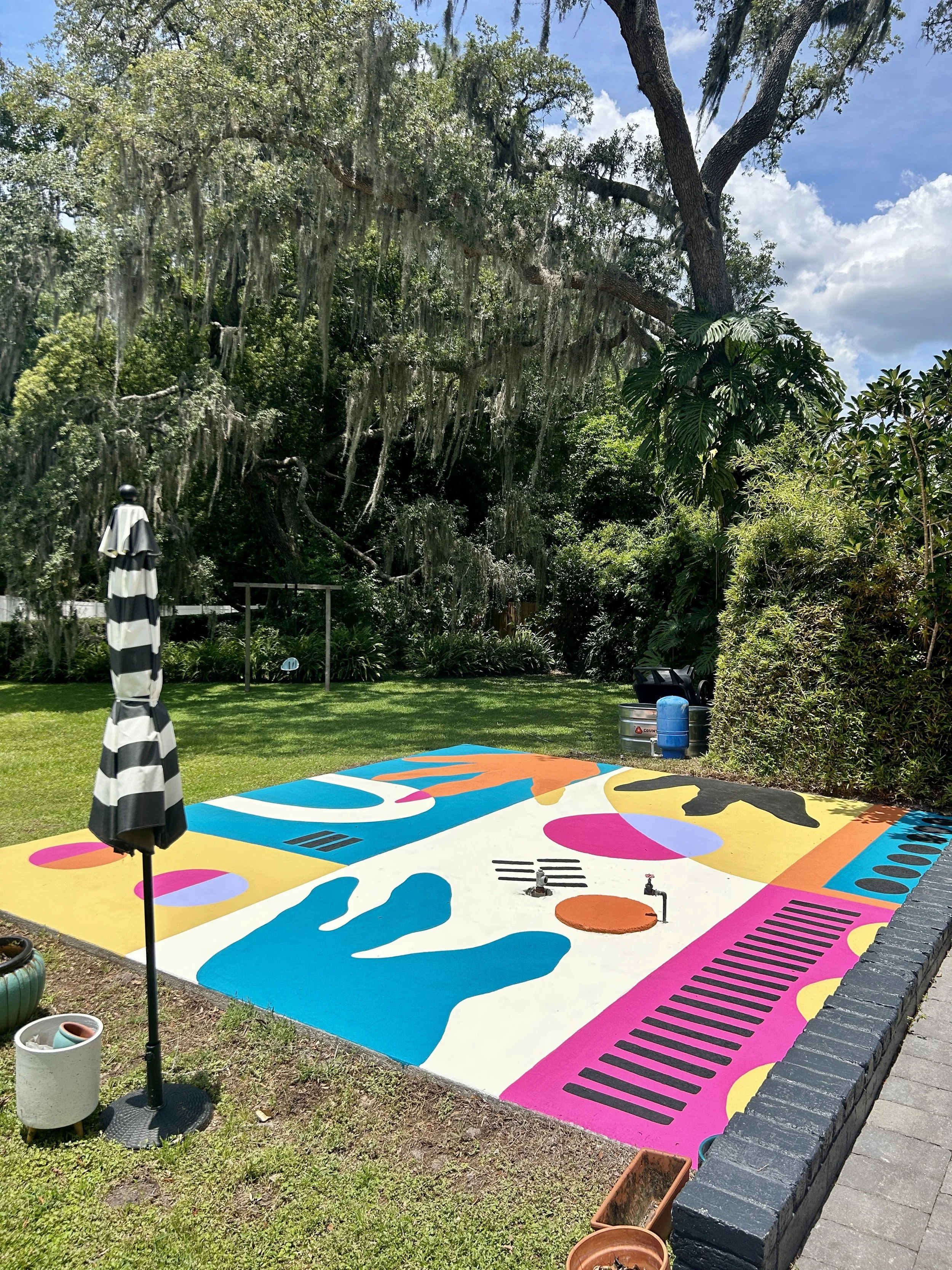 Colorful abstract mural painted on a section of concrete pavement in a backyard surrounded by greenery, trees, and outdoor furniture including a black and white striped umbrella.