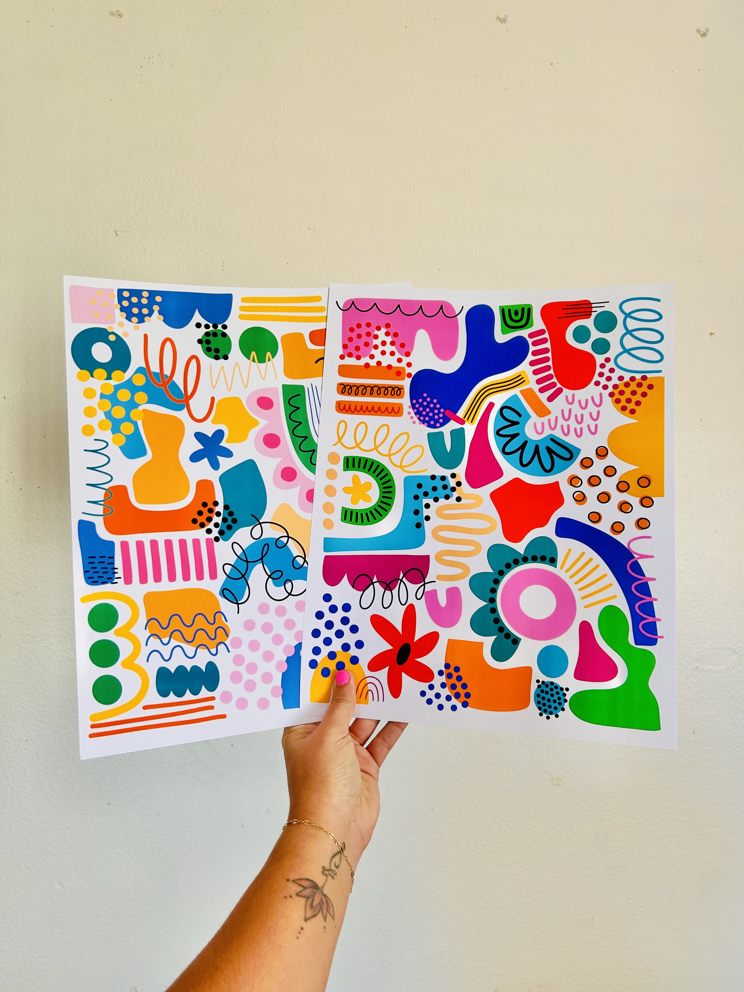 Brightly colored abstract doodle art on two sheets of paper held up against a plain wall.