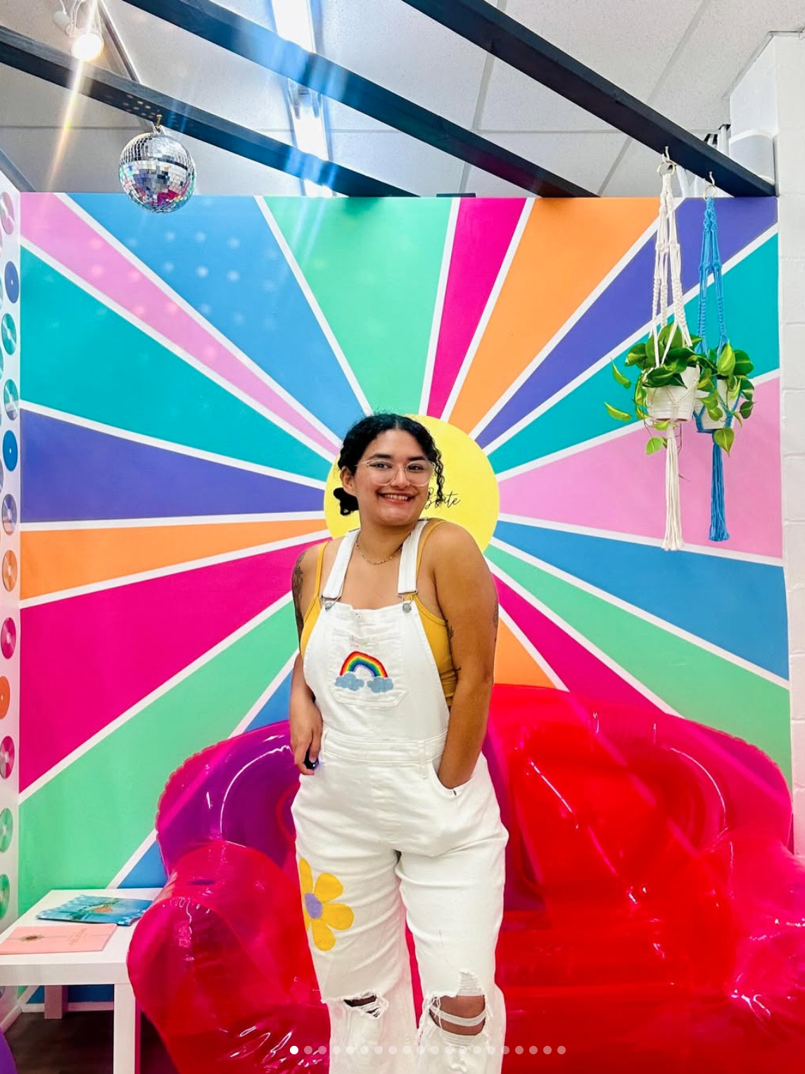 A woman with glasses and curly black hair, wearing a yellow tank top and white overalls with colorful patches, standing in front of a rainbow-colored wall with a sun design, smiling with her hands in her pockets, in a vibrant, fun indoor setting.
