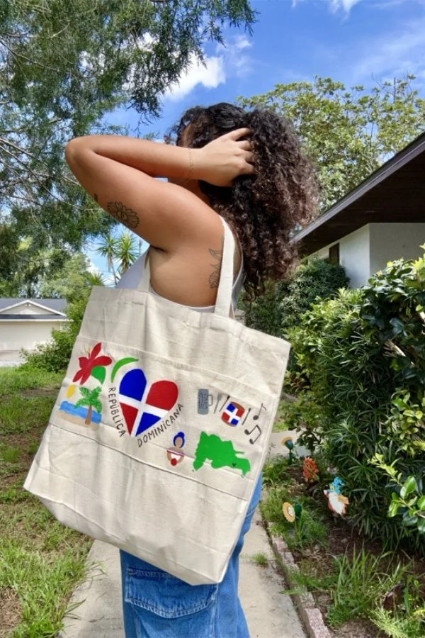 A woman with curly hair stands outdoors on a sunny day, holding her head with one hand. She carries a large tote bag decorated with colorful artwork representing the Dominican Republic, including a palm tree, beach, and musical notes. She is wearing a beige tank top and blue jeans, with green foliage and a house in the background.