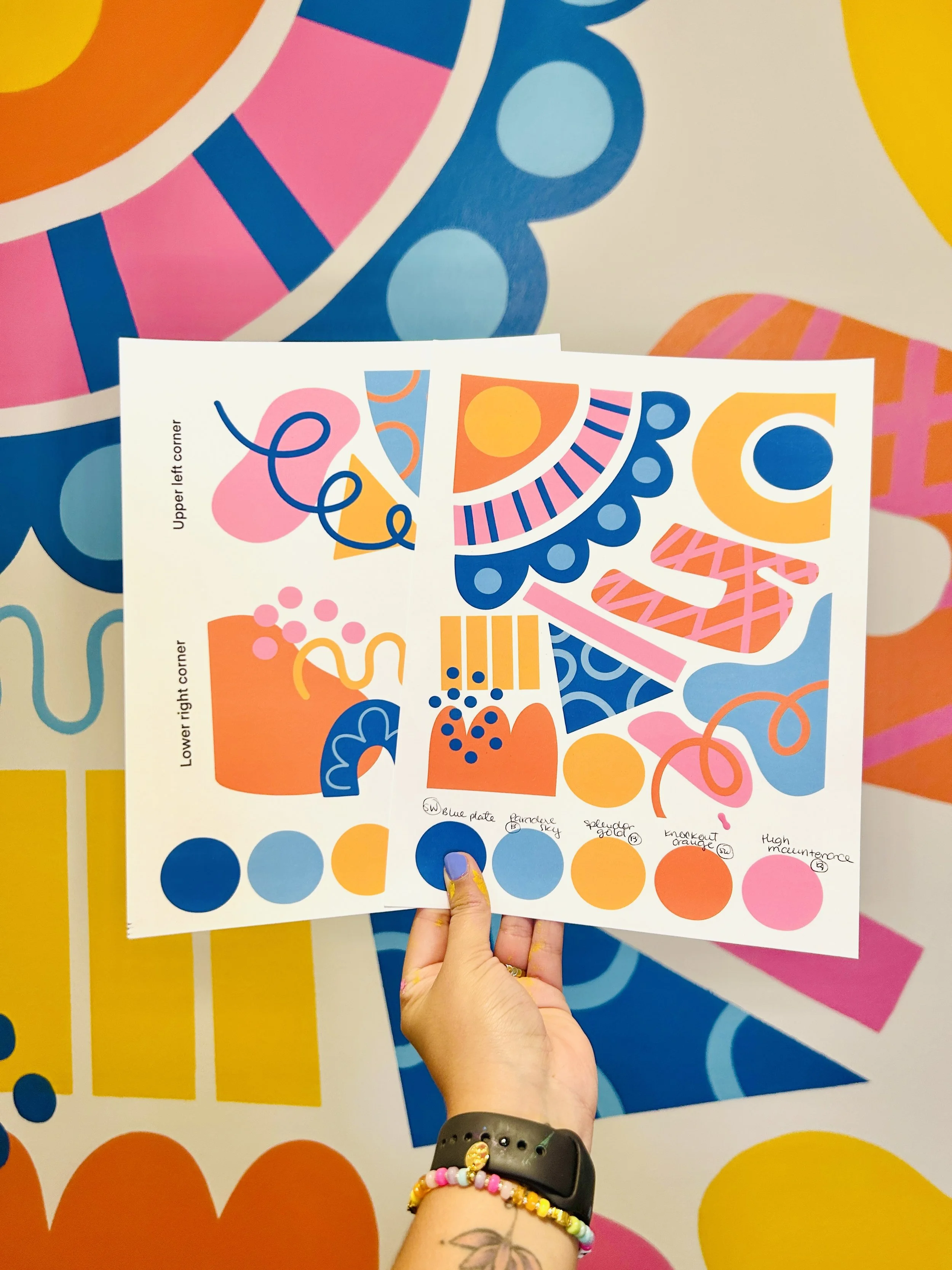 A person holding two colorful sheets of abstract, geometric designs and patterns, with a background of similar abstract shapes and patterns, featuring bright colors such as pink, blue, orange, and yellow.