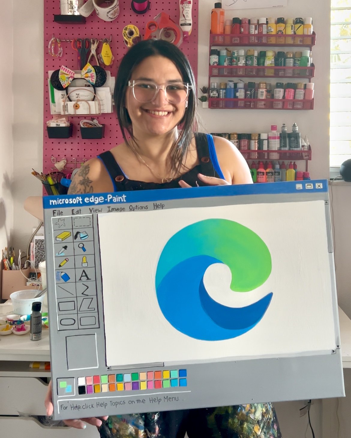 A woman smiling and holding a computer screen showing the Microsoft Edge Paint logo, in a craft room with shelves of paints, art supplies, and a pink pegboard with hanging tools and accessories.
