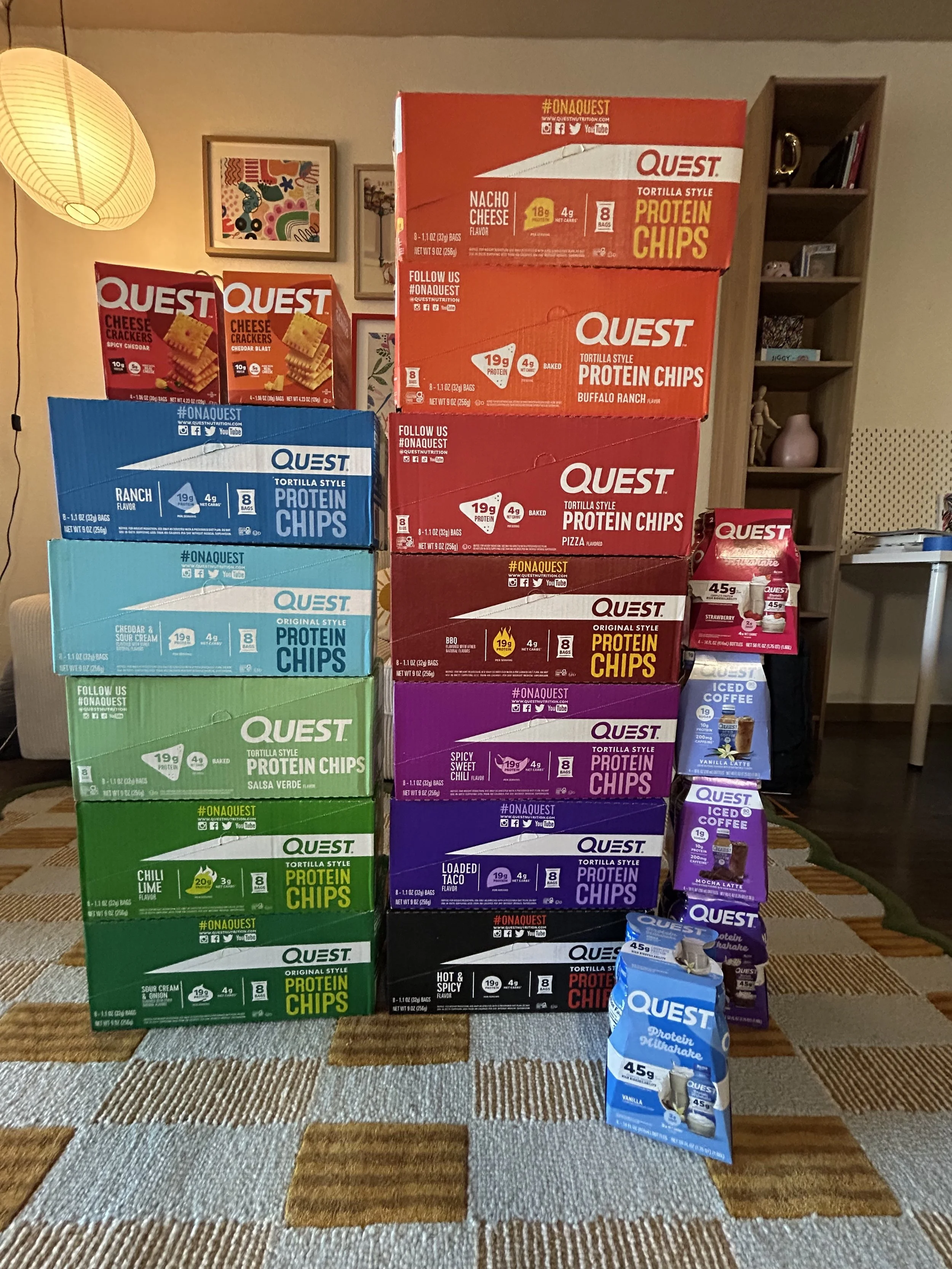 A stack of Quest protein chip boxes and smaller Quest snack packs on a carpeted floor in a living room, with framed pictures, shelves, and a lamp in the background.