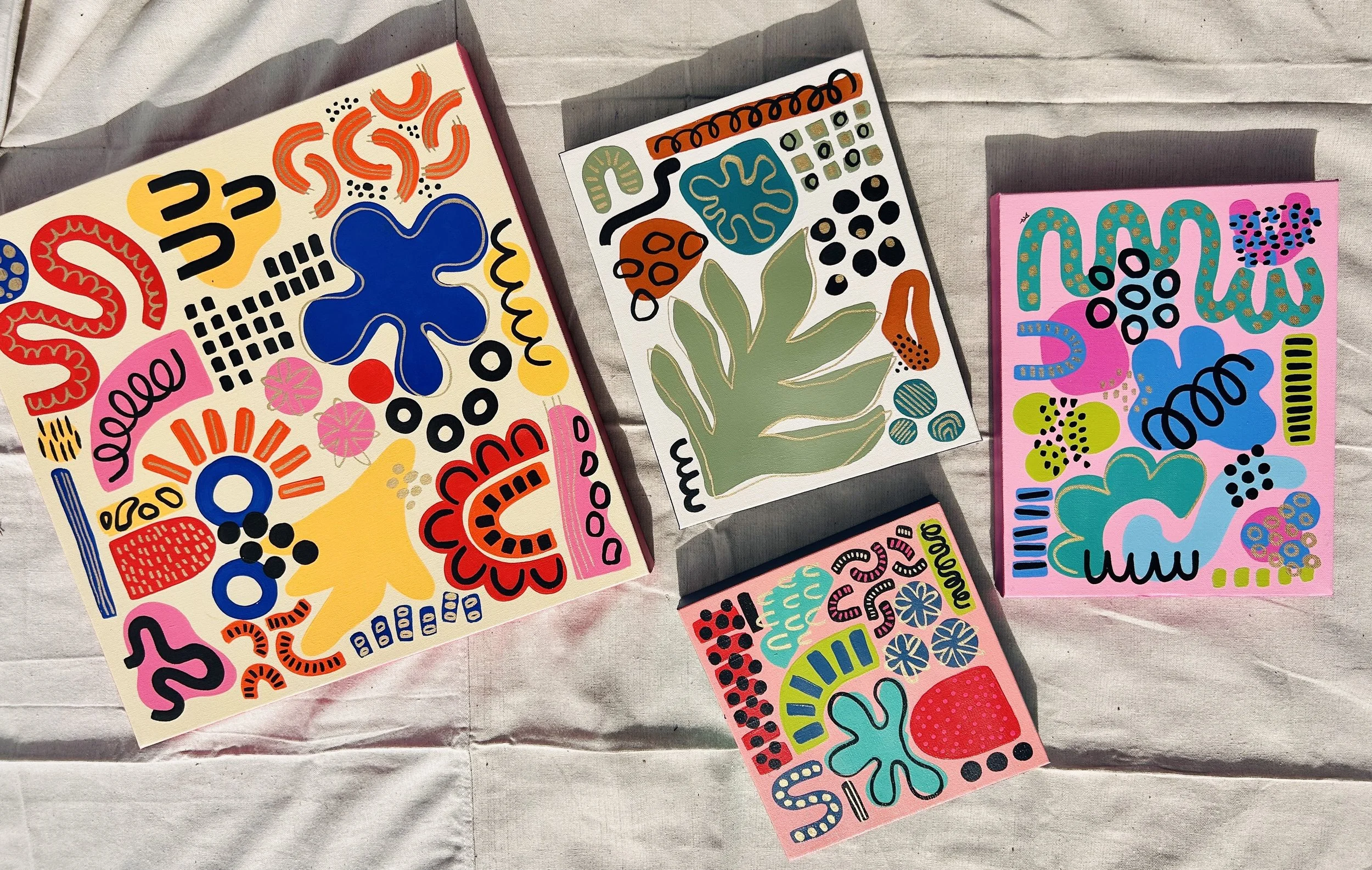 Four colorful abstract art pieces with various shapes and patterns laid out on a fabric surface.
